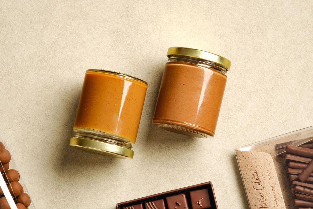 Two glass jars filled with caramel spread beside a chocolate bar on a beige surface.
