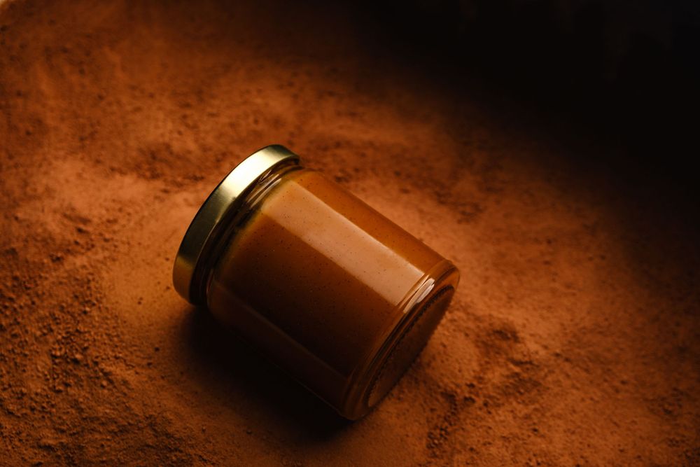 A glass jar of caramel spread lying on its side in cocoa powder under warm light.