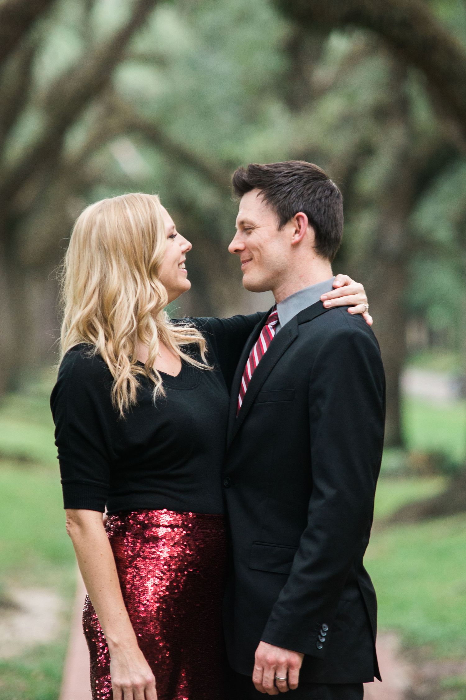 Couple embracing and laughing in formal wear with a sparkly red sequin dress against a natural backdrop.