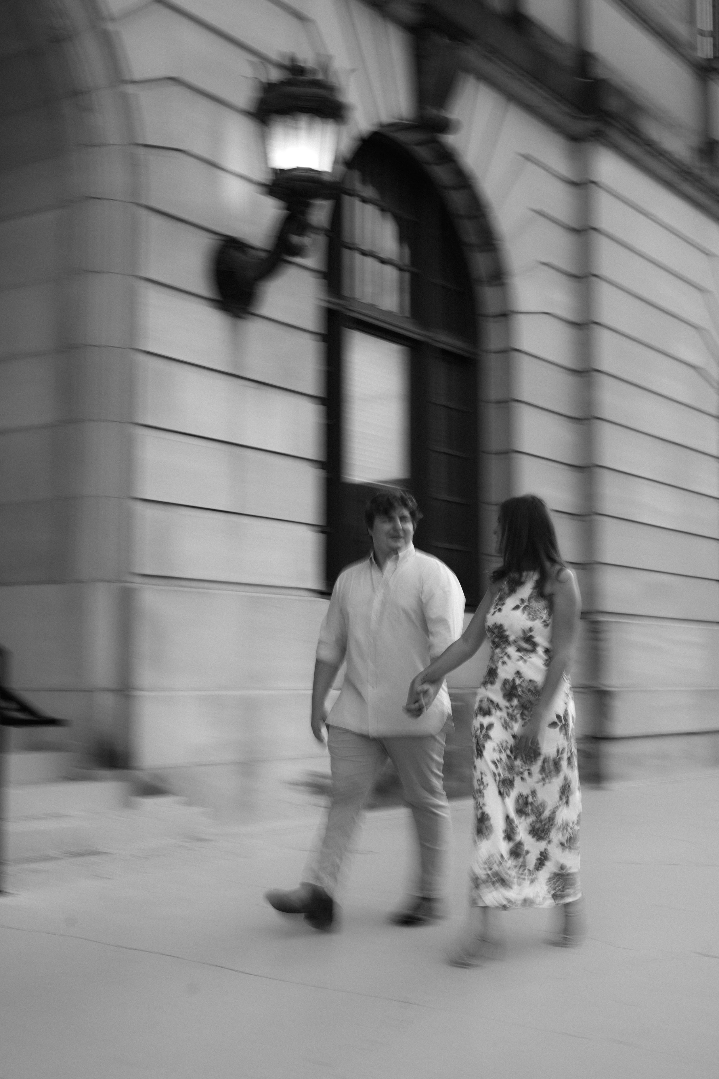 Motion blur black and white photo of couple walking along stone building exterior.