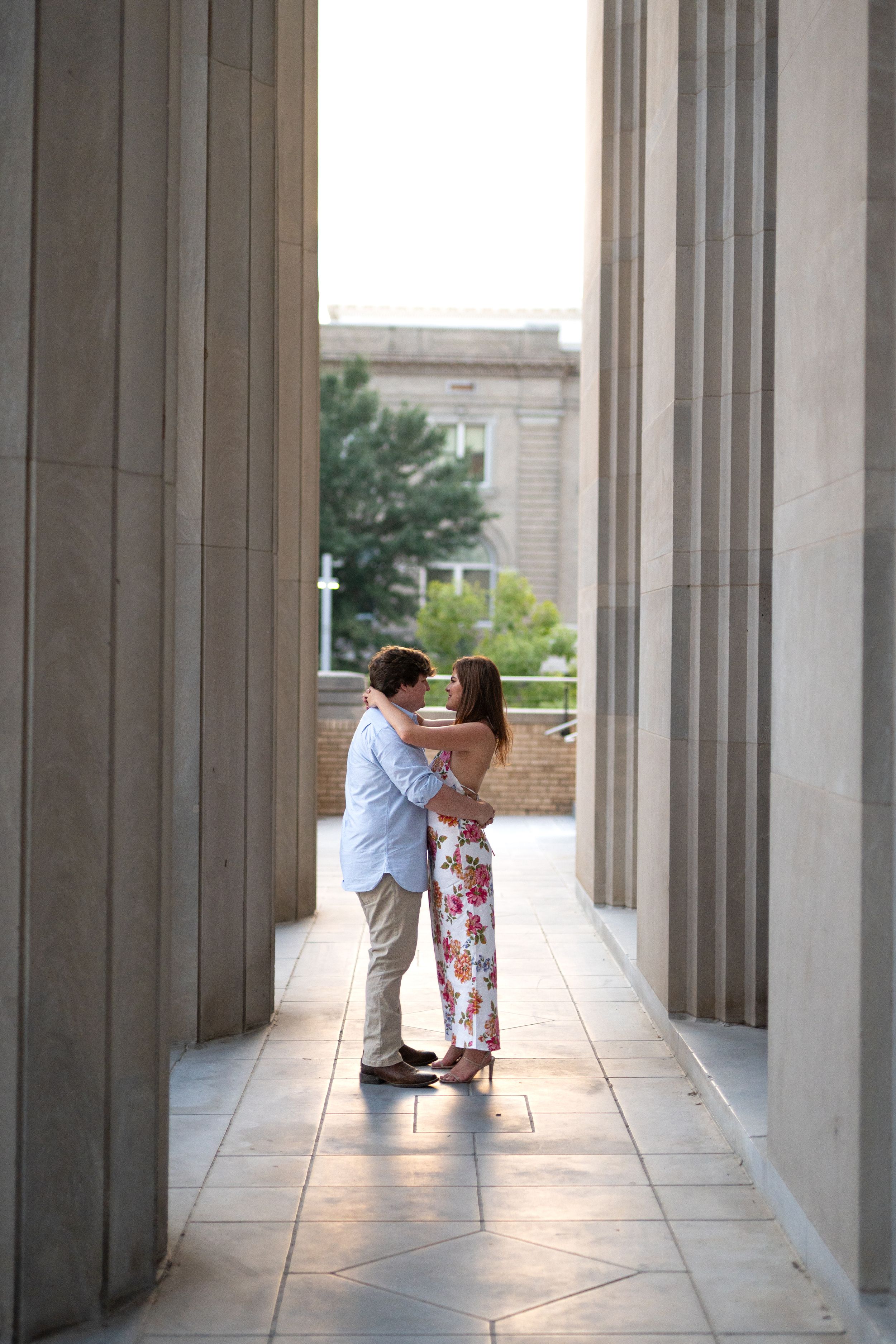 A couple shares an intimate moment in a columned hallway with natural light streaming through.