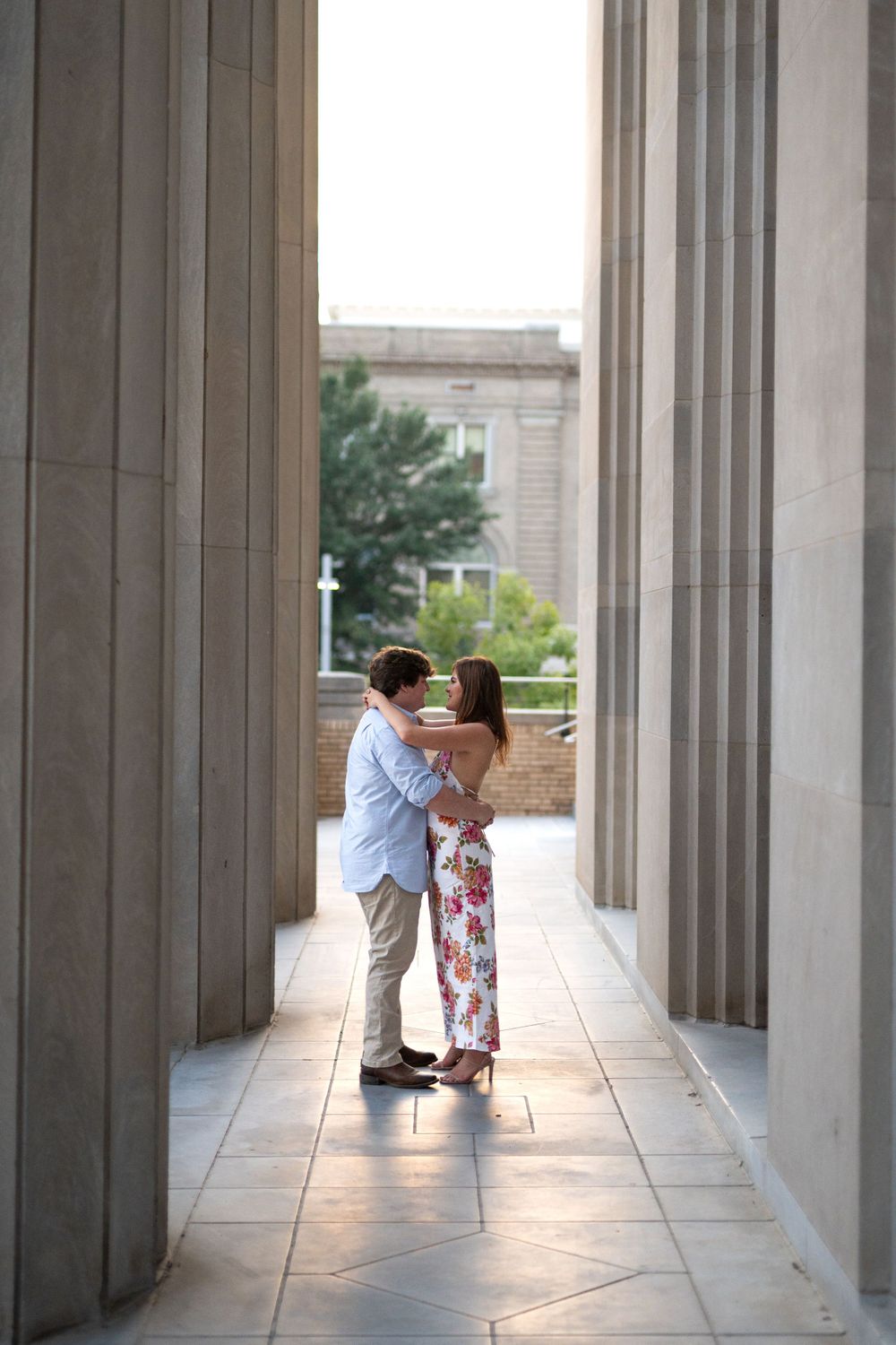 A couple shares an intimate moment in a columned hallway with natural light streaming through.