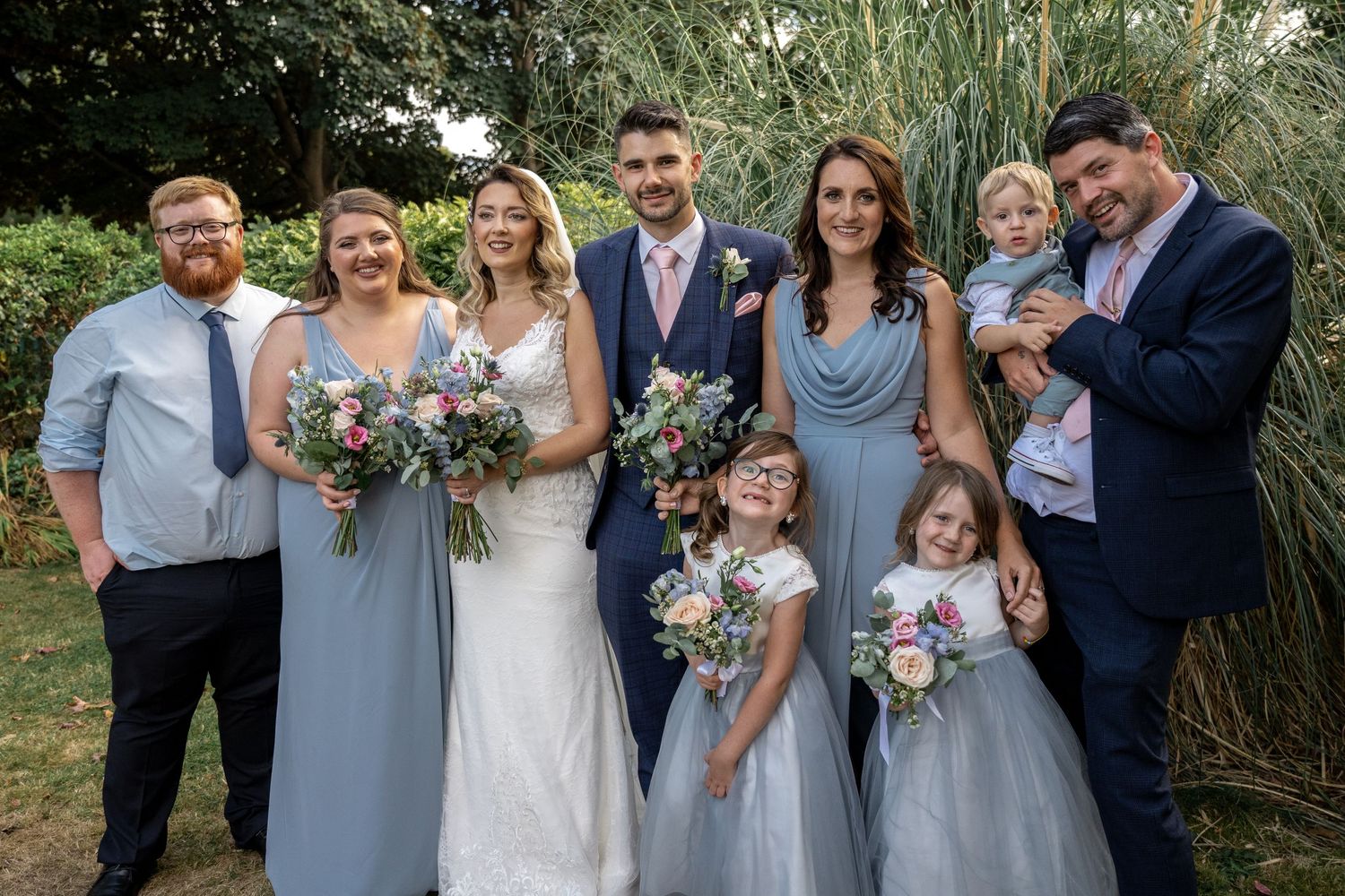 Wedding party group photo with bridesmaids in light blue dresses and children in formal attire outdoors.