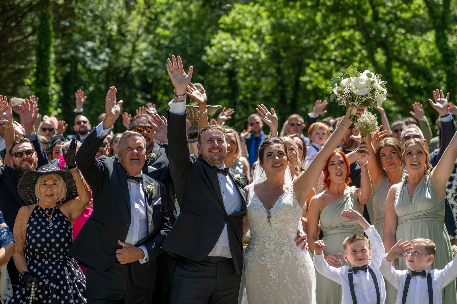 Wedding group photo with guests raising hands in celebration under green trees.