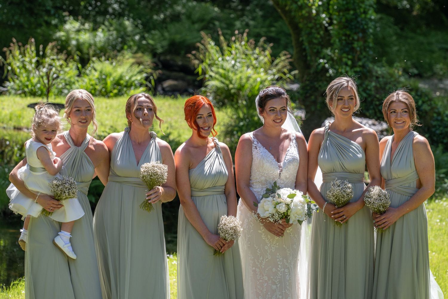 Bridesmaids in sage green dresses holding bouquets pose together in sunny garden setting.