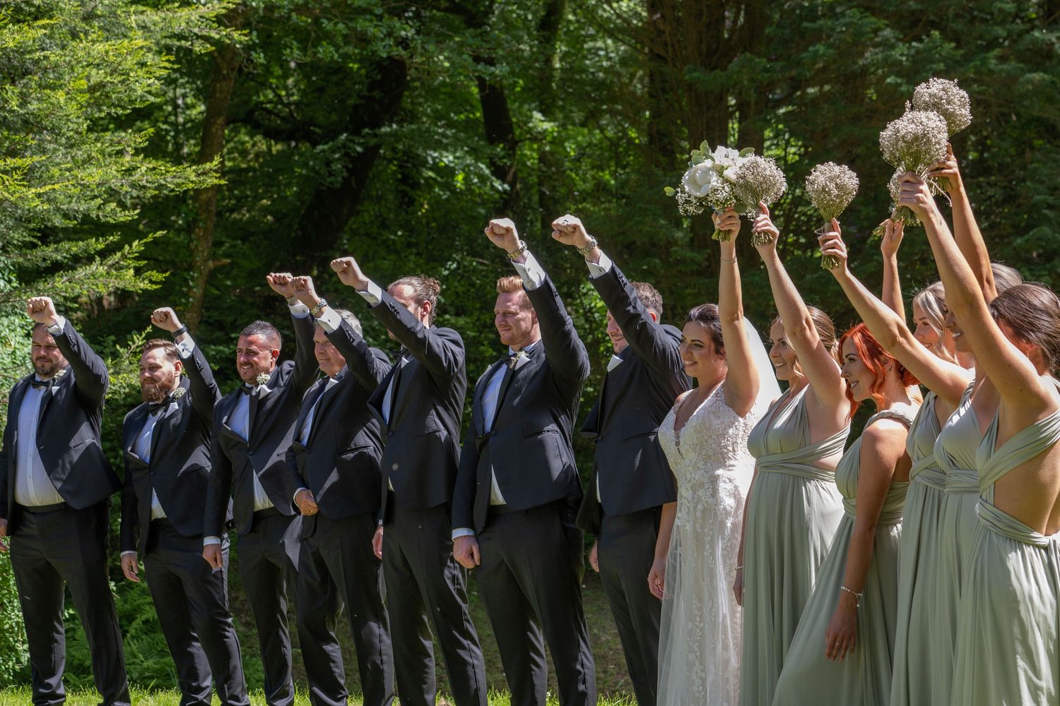 Wedding party standing in a line outdoors raising bouquets and fists in celebration with trees in background.