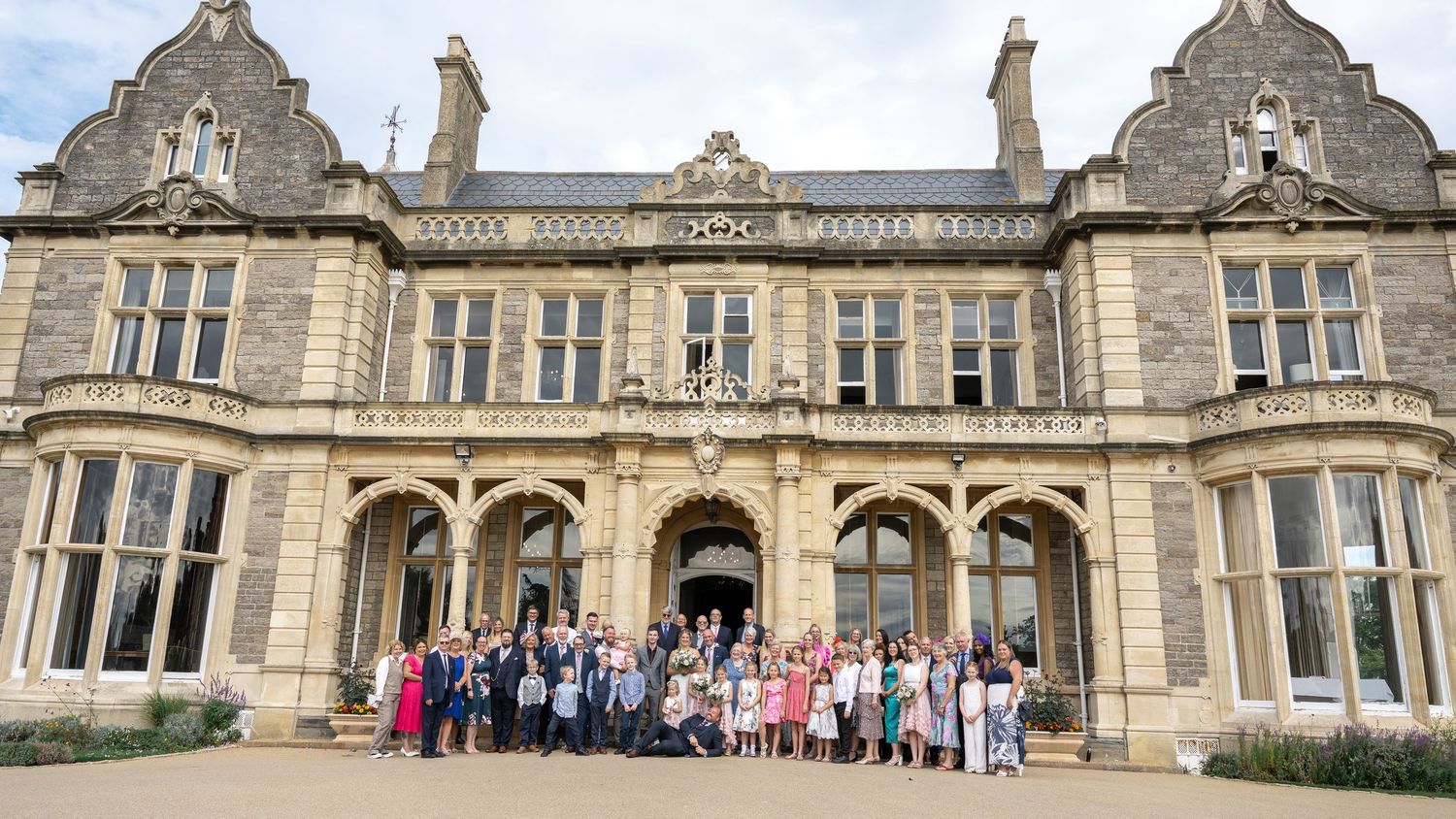 Wide view of wedding party and guests gathered in front of majestic Victorian stone mansion.