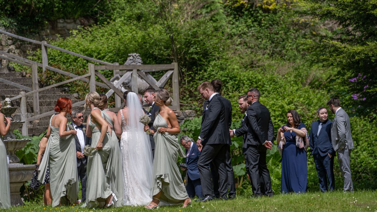 Wedding group gathered on grassy hillside for informal photos near rustic wooden fence.