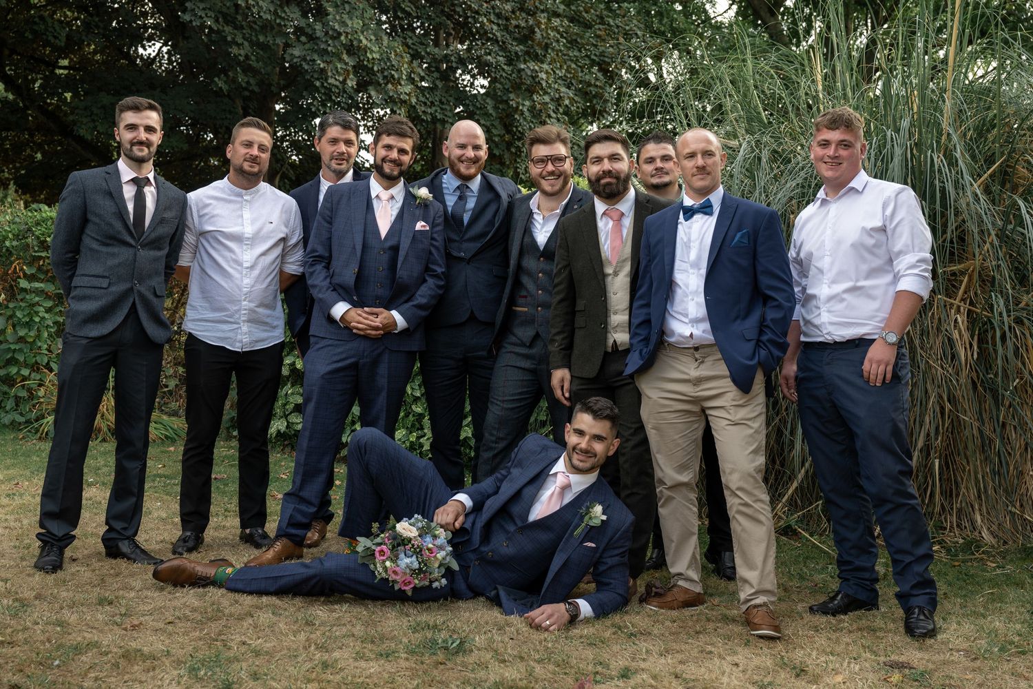 Groomsmen in suits posing casually for group photo on grass with tall plants in background.