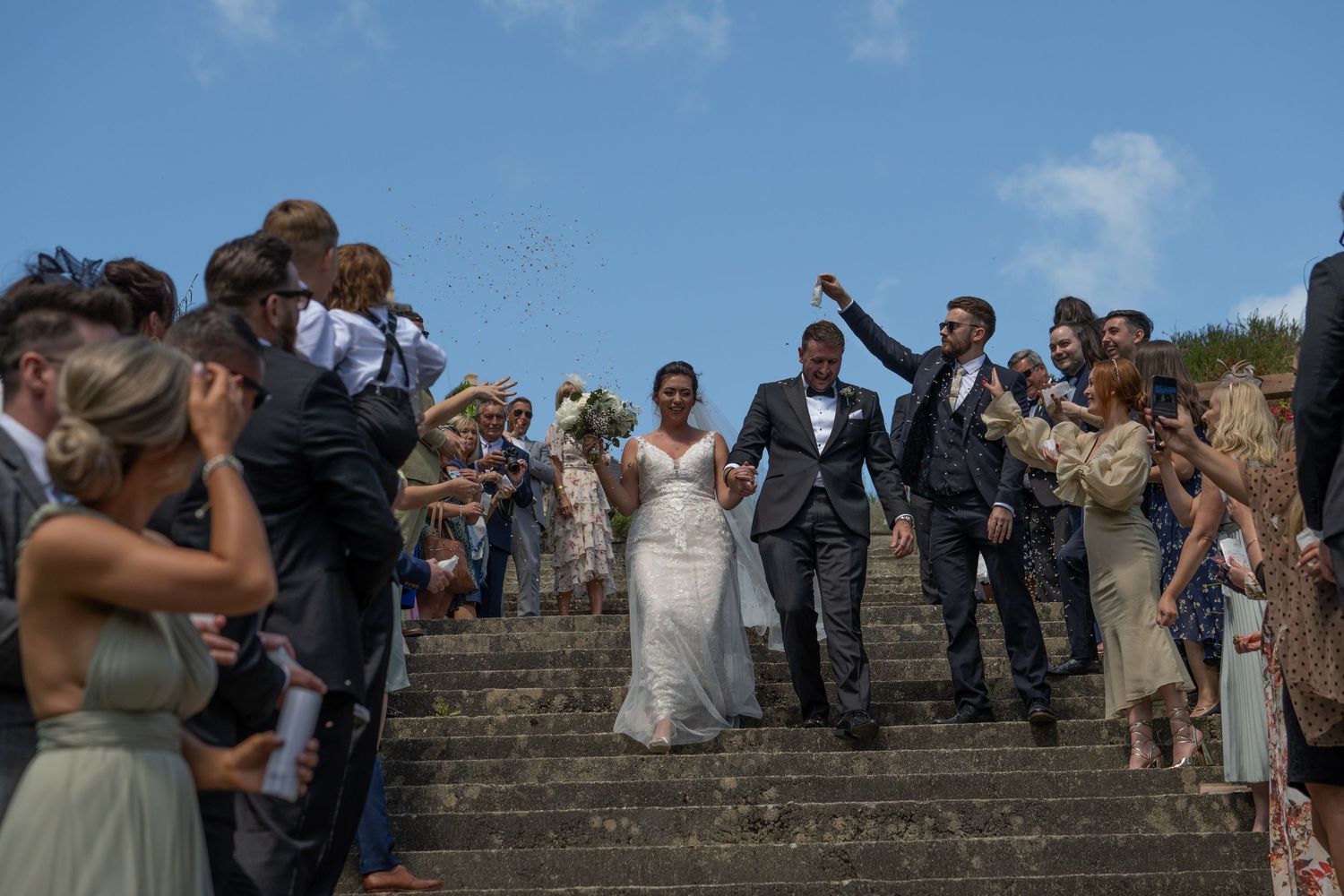Wedding party celebrating on grand stone steps with confetti flying through the air.