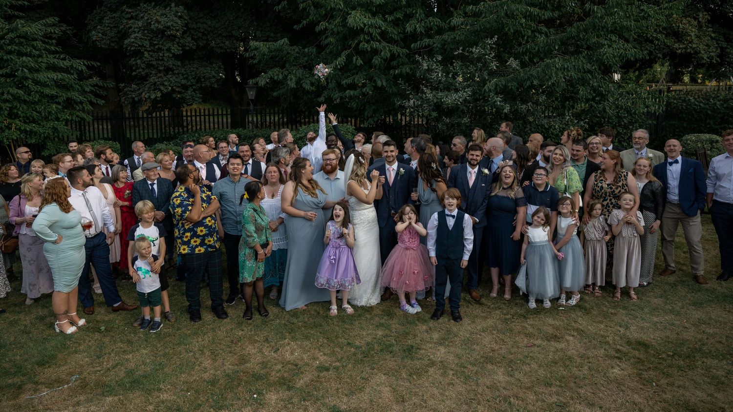 Large wedding group photo of guests and wedding party on lawn with trees in background.