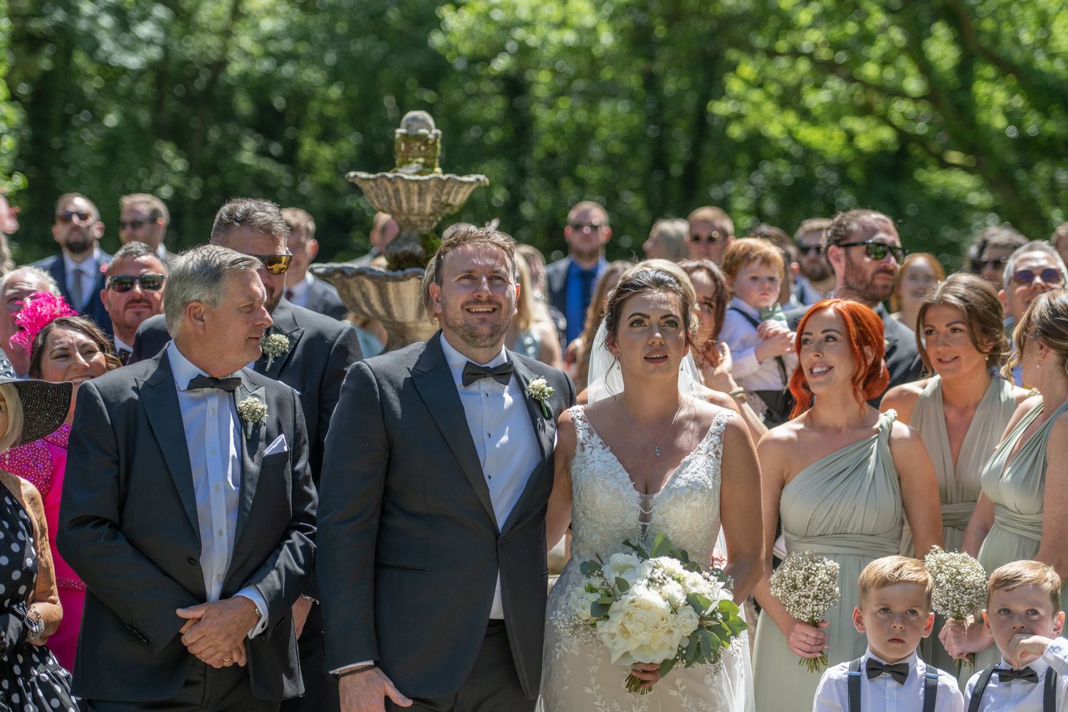 Wedding party and guests gathered outdoors for group photos on a sunny summer day.