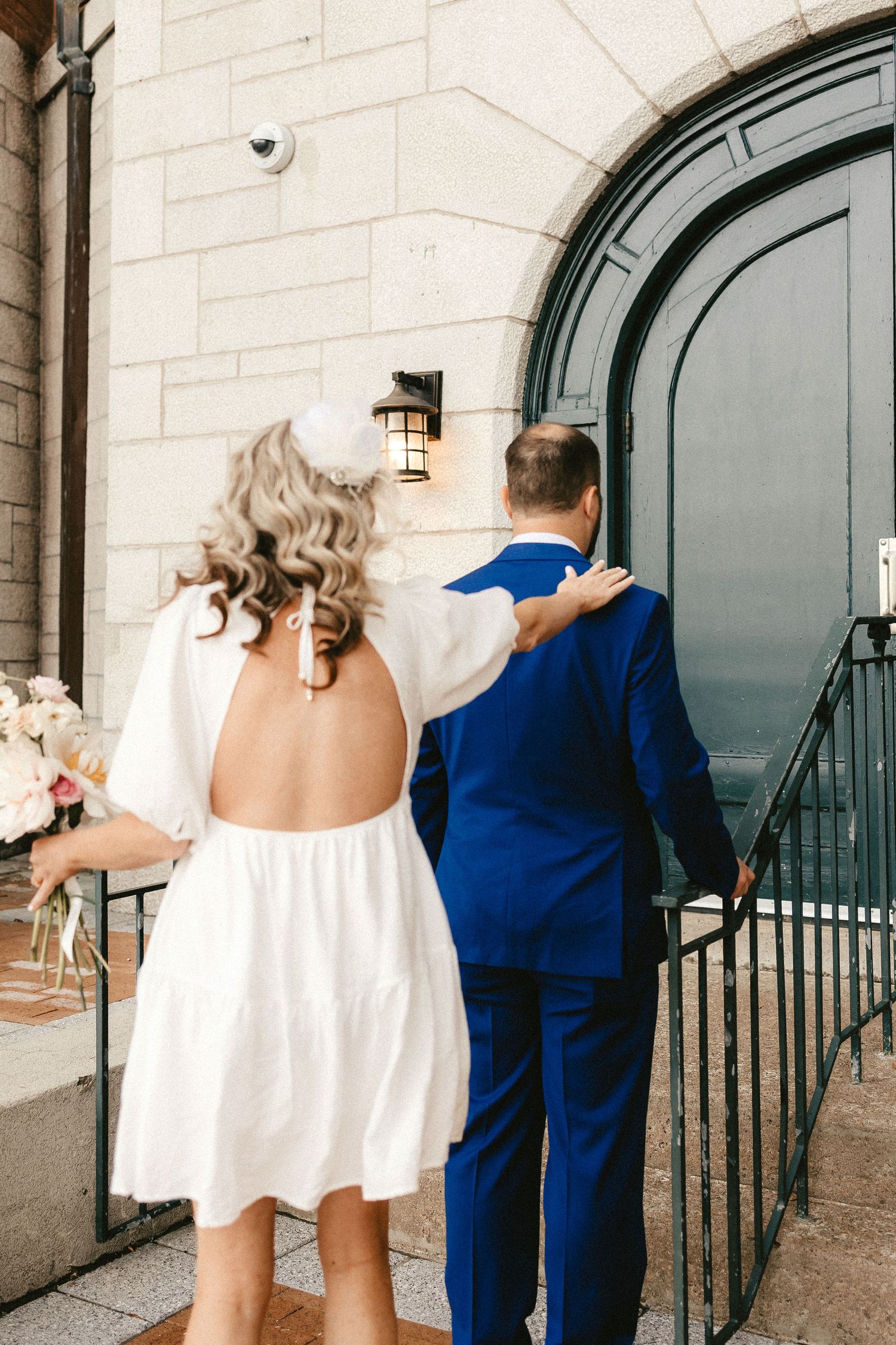 A backless white wedding dress and blue suit shown from behind against an arched doorway.
