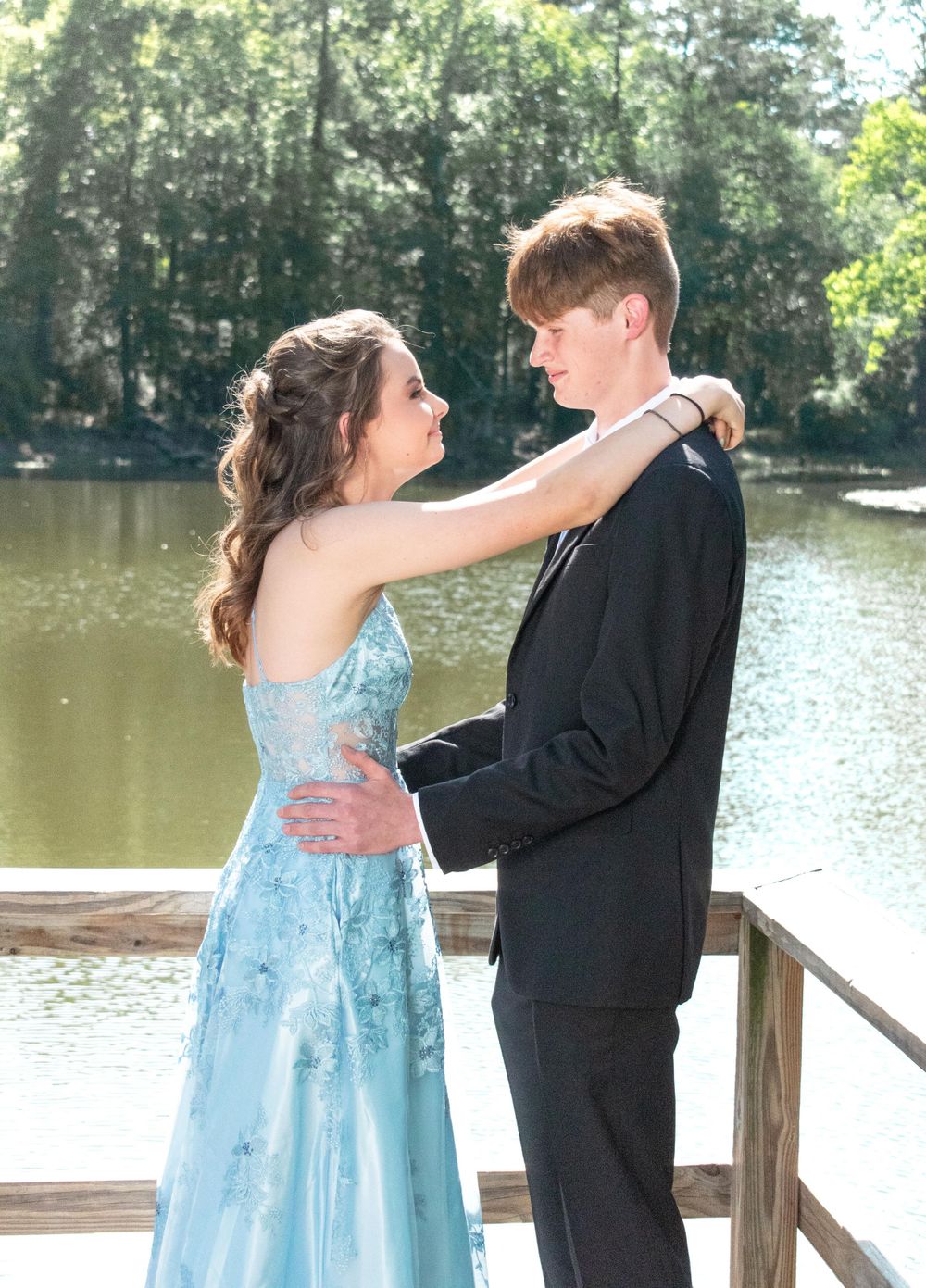 A couple in formal wear share an intimate moment on a wooden dock by a serene lake.