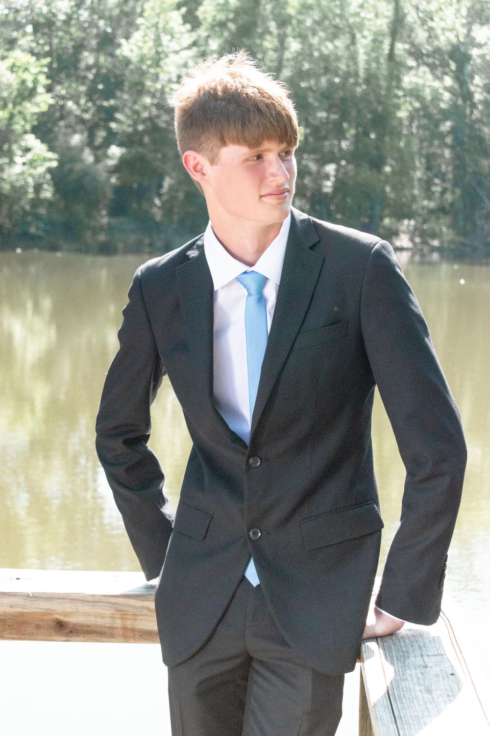 Young man in classic black suit and blue tie poses by lake.