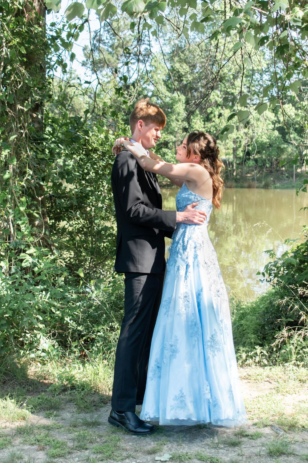 A couple in formal attire embrace by a lake, with the woman wearing a flowing light blue gown.