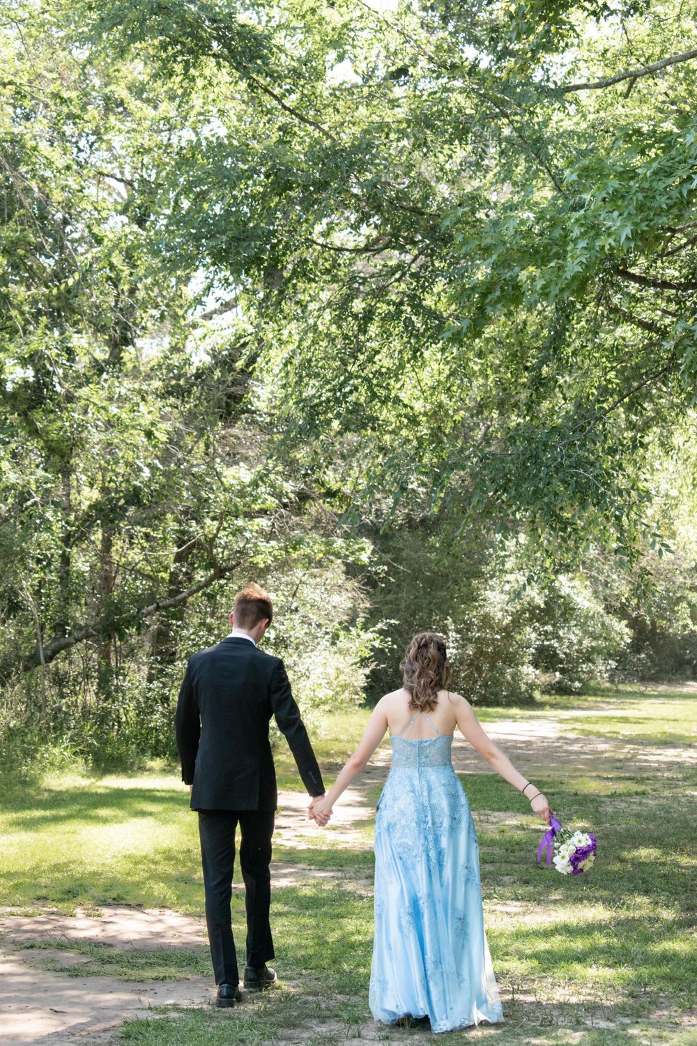 A couple walks hand in hand along a path surrounded by trees, with the woman in a blue prom dress.