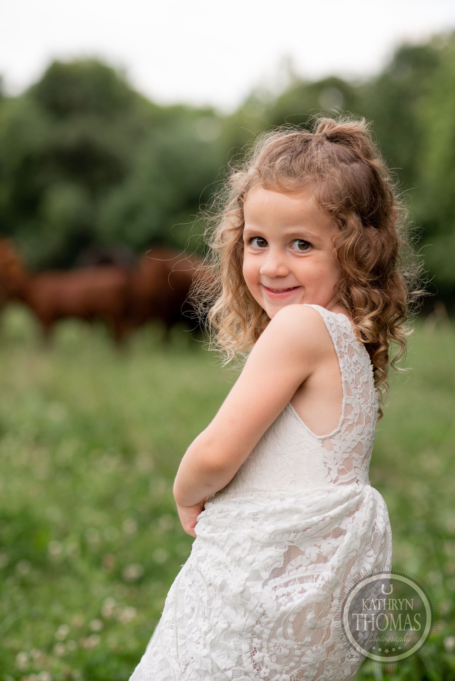 A young person in a white lace dress poses playfully in a field with red cattle grazing behind.