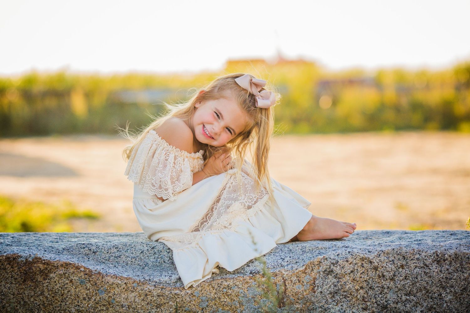 Young girl in white dress laughing playfully while sitting on a rock at golden hour.