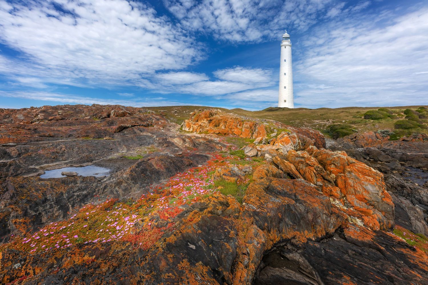 White lighthouse stands tall on colorful rocky terrain against vibrant blue sky with scattered clouds.
