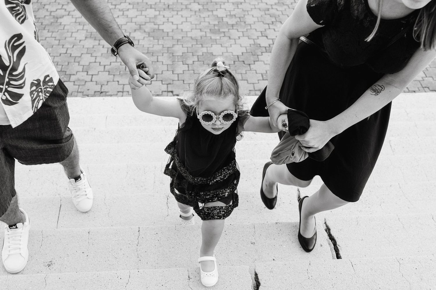 A small child in sunglasses jumps between two adults on a paved surface.