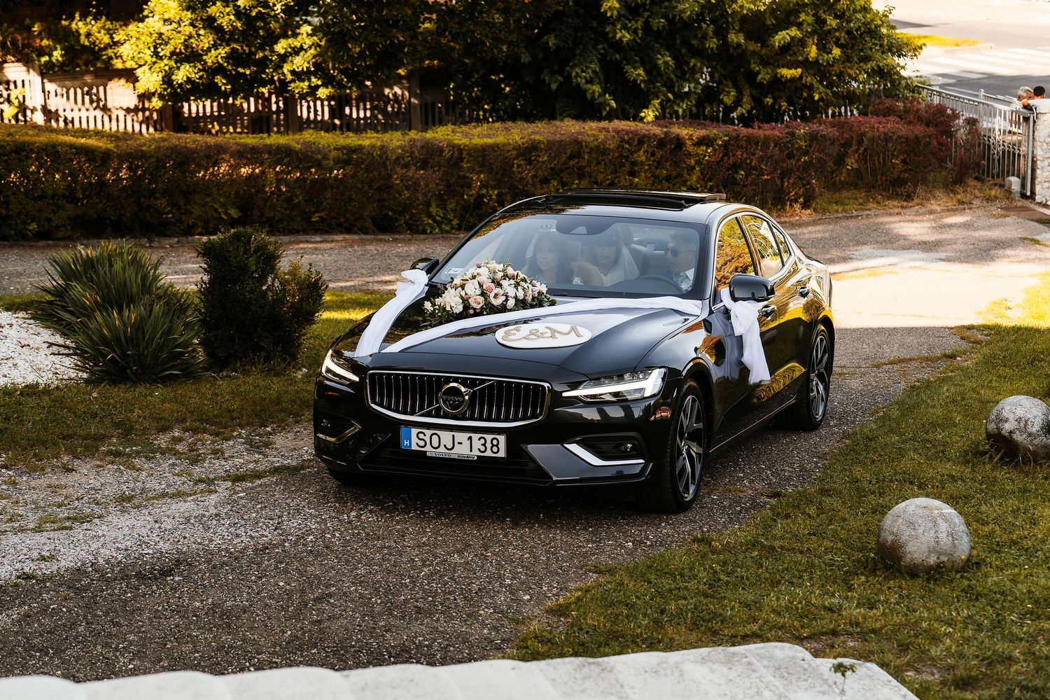 A black Volvo sedan decorated with wedding flowers parked in a scenic garden driveway.