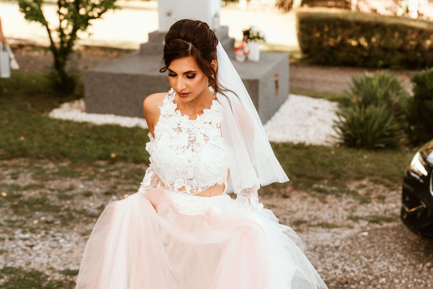 A person in an elegant white lace wedding dress sits outdoors during sunset.