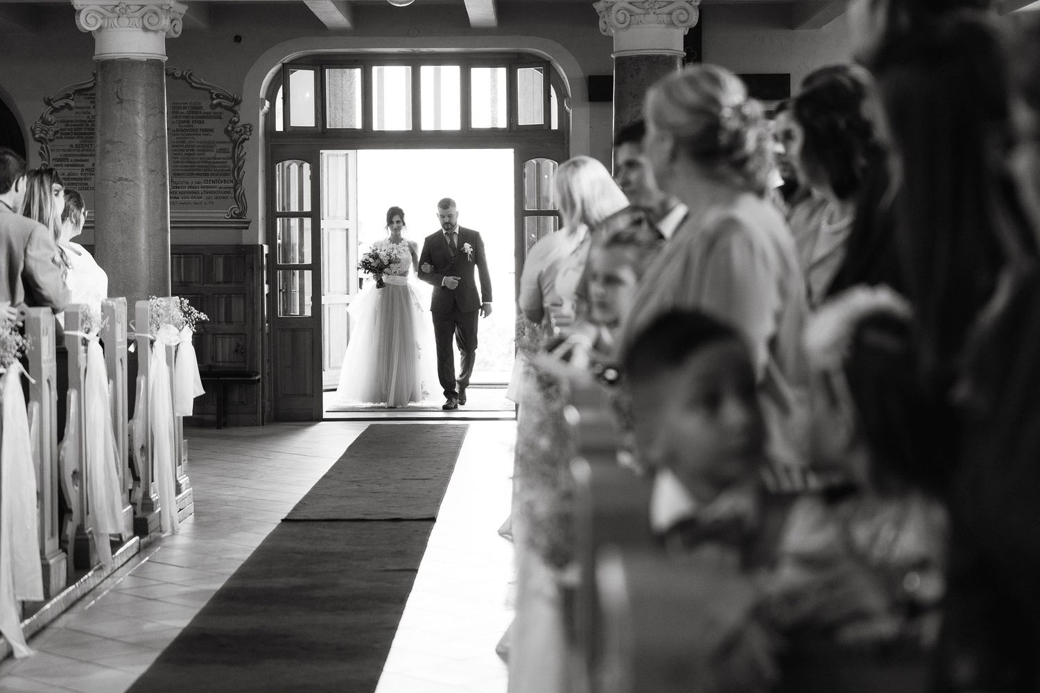 A black and white view down a church aisle filled with wedding guests watching the ceremony.