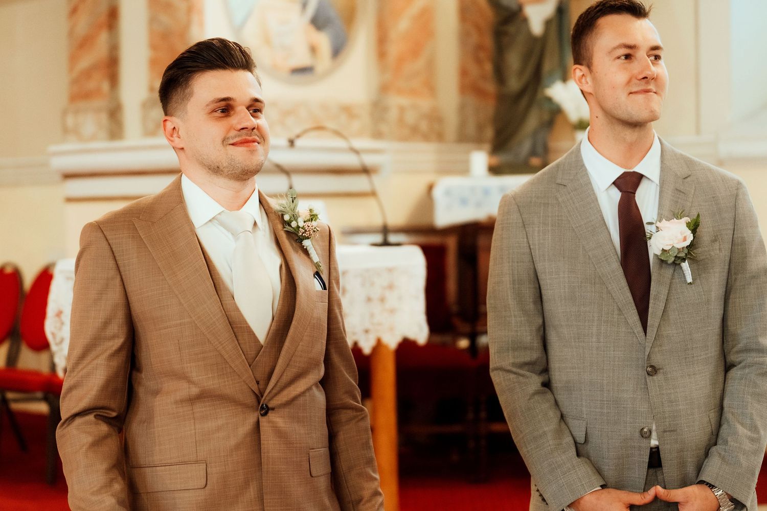 Two groomsmen wearing suits, one in tan and one in gray, stand at the altar during a wedding ceremony.