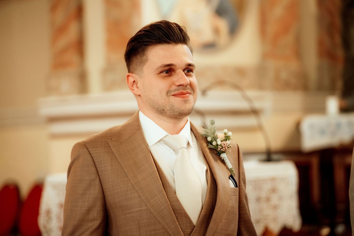 A groom in a tan suit smiles during his wedding ceremony in an ornate church setting.