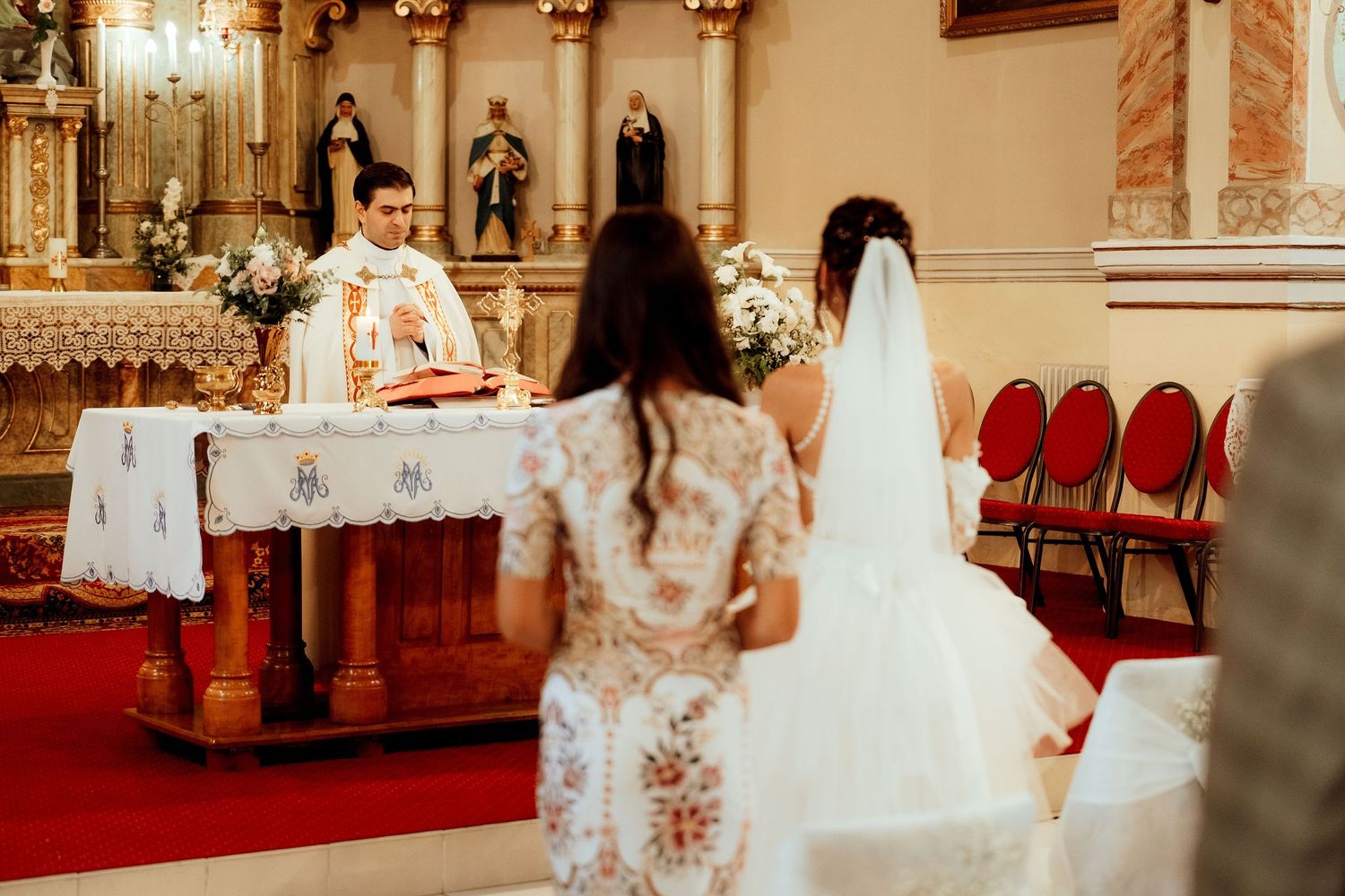 A wedding ceremony in progress at an ornate Catholic church with red chairs and floral decorations.
