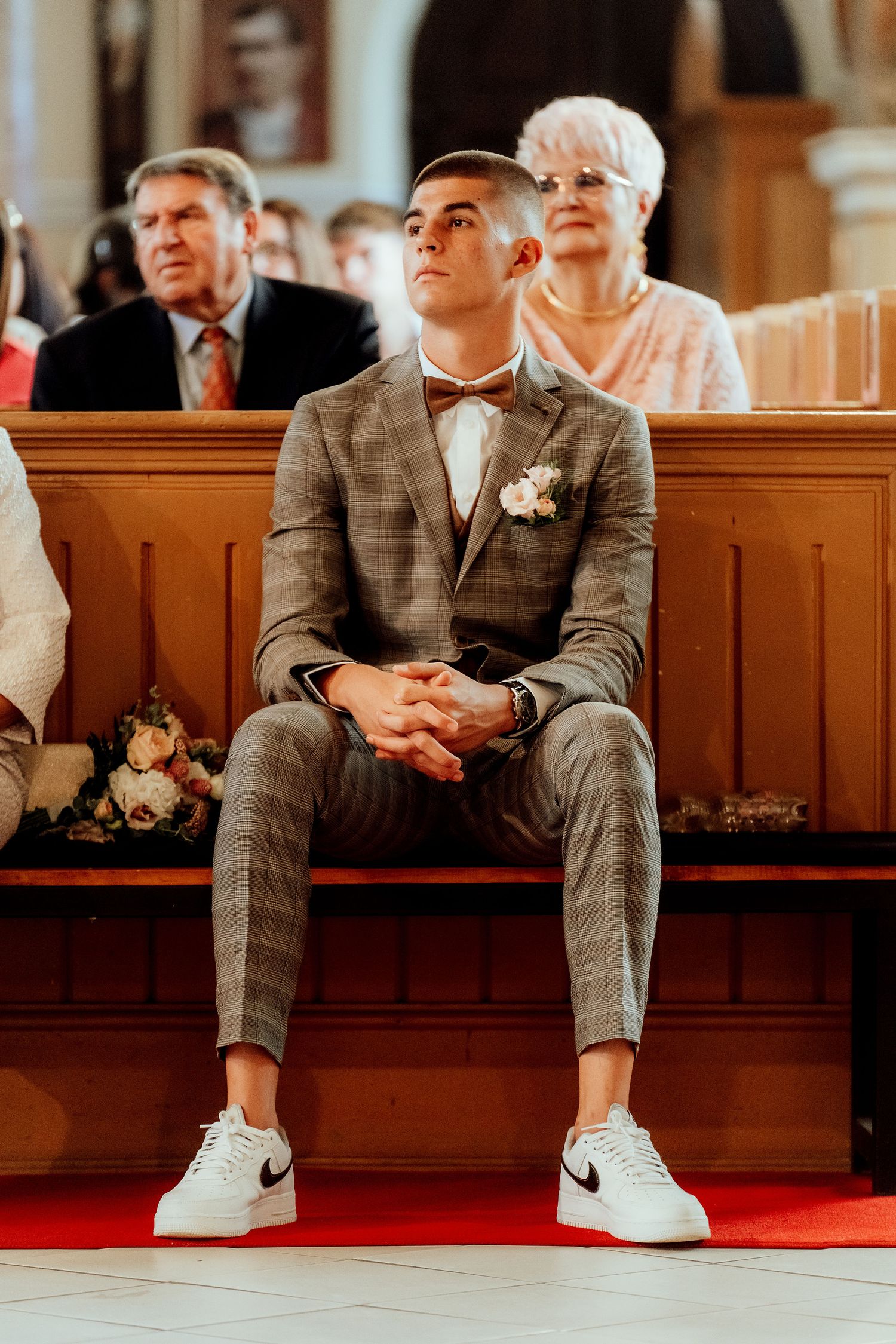 Modern groom in plaid suit and Nike sneakers sits in church pew during ceremony.