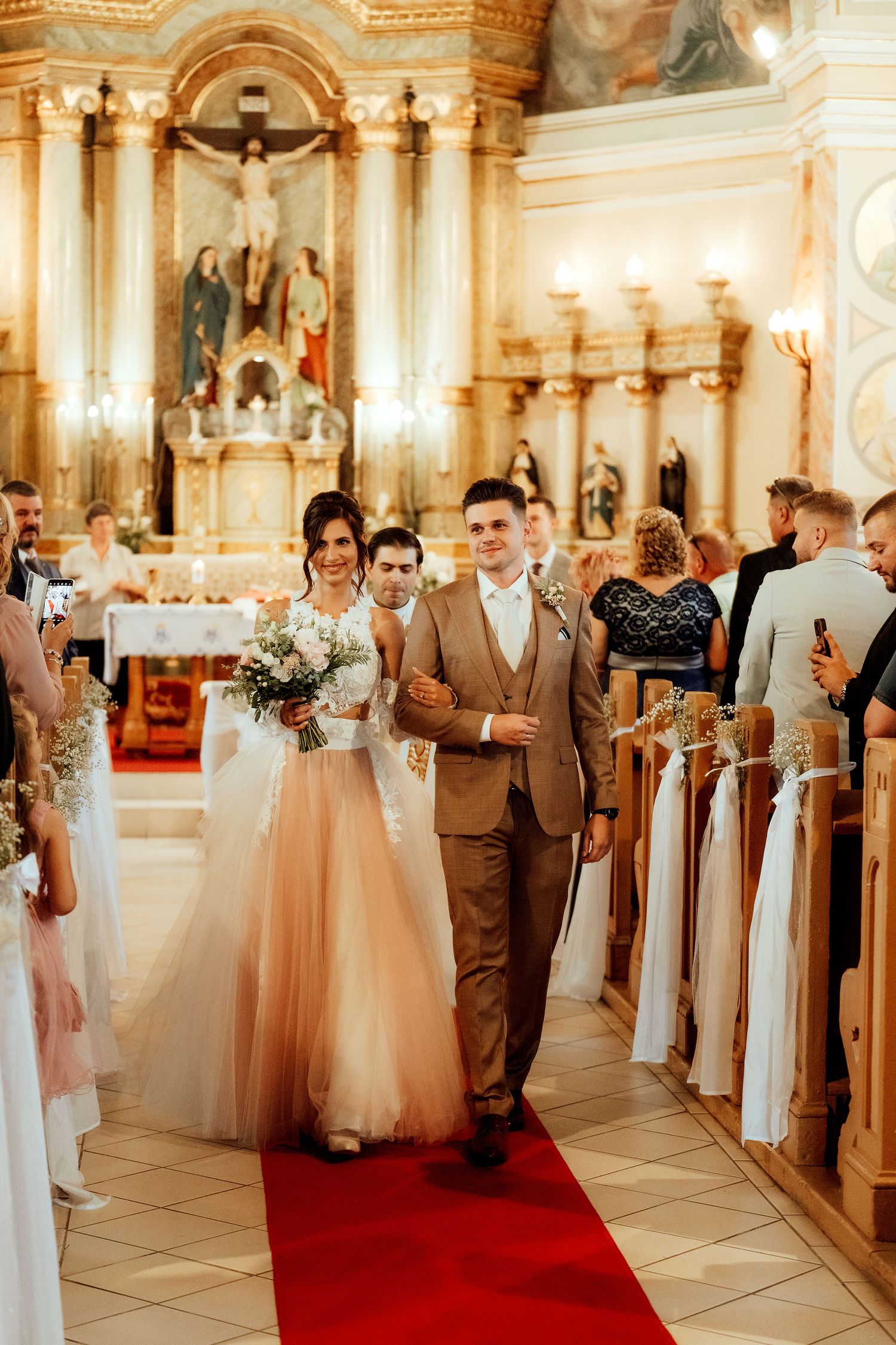 Wedding ceremony in a grand cathedral with red carpet aisle and white pew decorations.