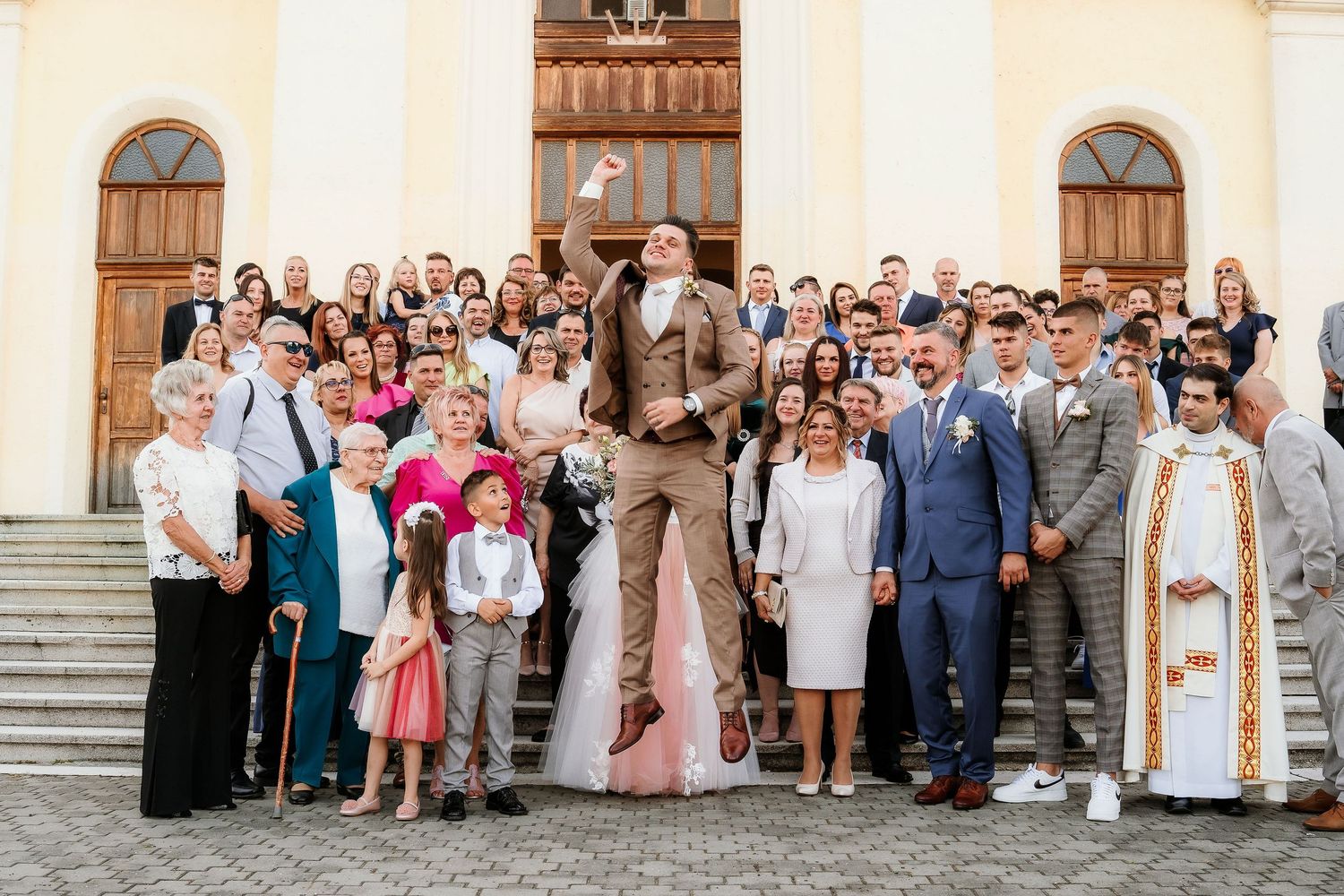 Large wedding party group photo on church steps with guests in formal attire.