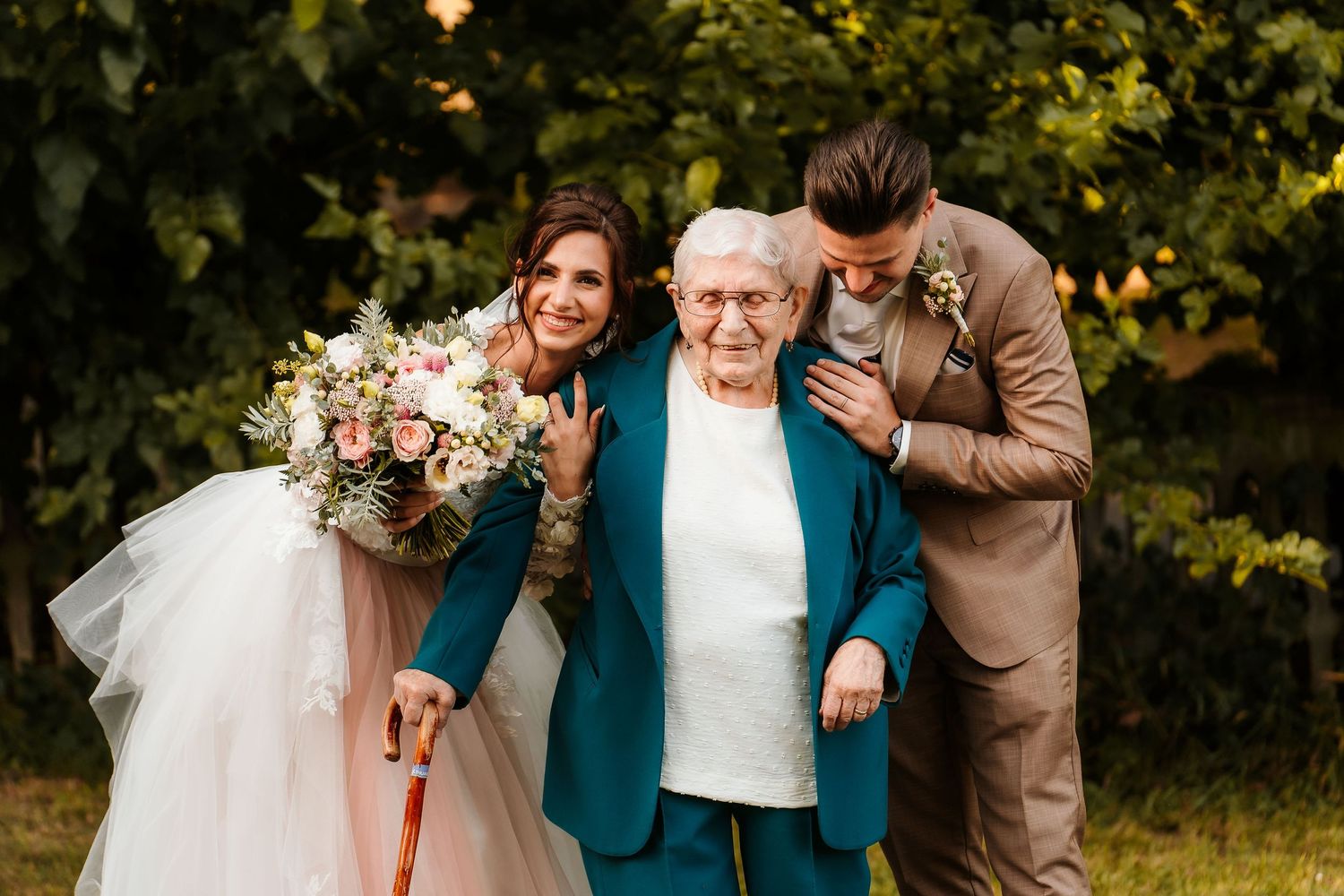 Two wedding guests helping an elderly woman in a blue jacket walk outdoors.