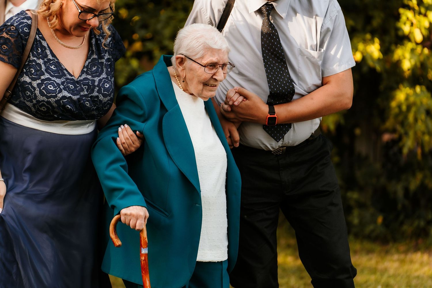 Family members assisting an elderly relative in a turquoise jacket during outdoor event.