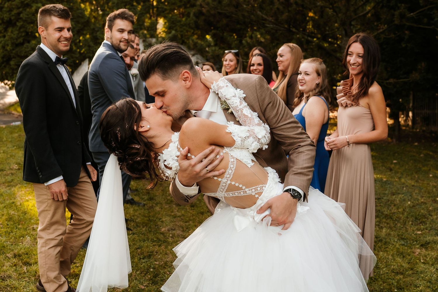 Romantic dip kiss moment between newlyweds surrounded by wedding party outdoors.