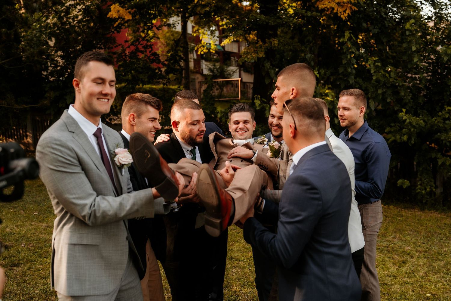 Groomsmen in suits share laughs while gathered in a circle outdoors during autumn.