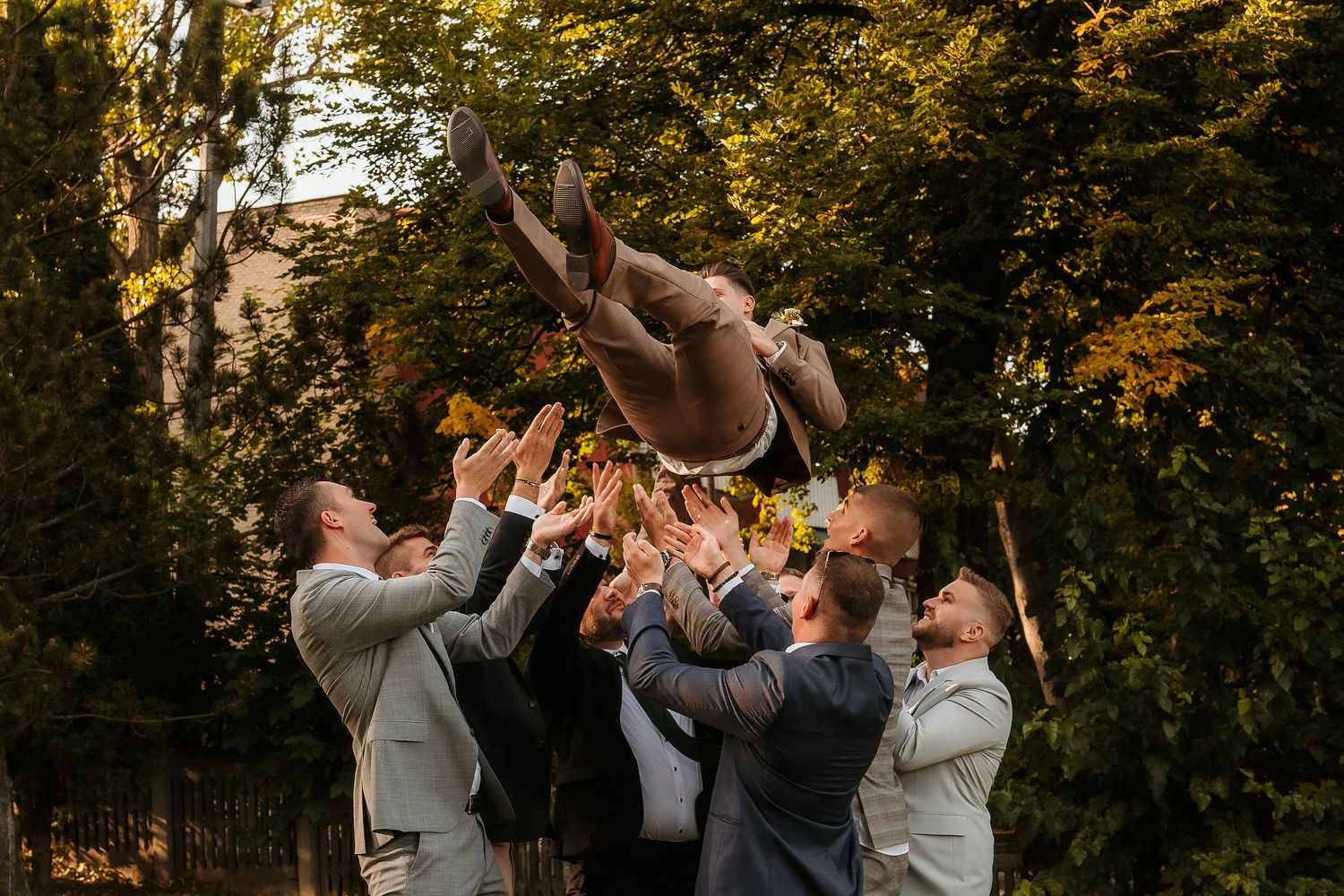 Wedding party throws someone into air during fun outdoor photo session with fall foliage.