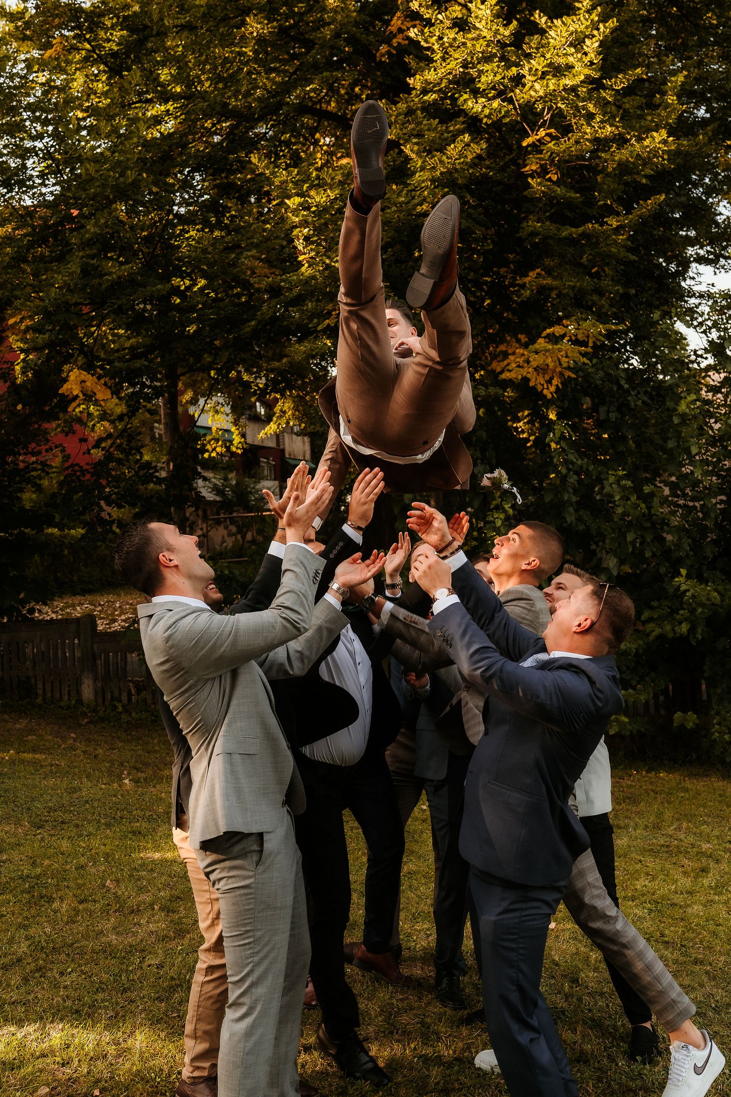 Group tosses person high into air during playful outdoor celebration with autumn trees.