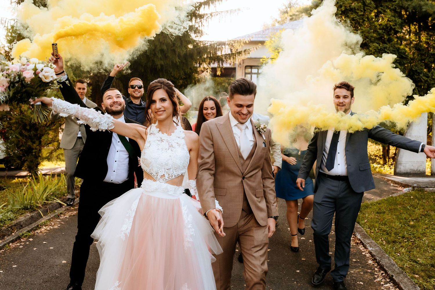 Wedding party walks down path with yellow smoke effects during outdoor celebration.