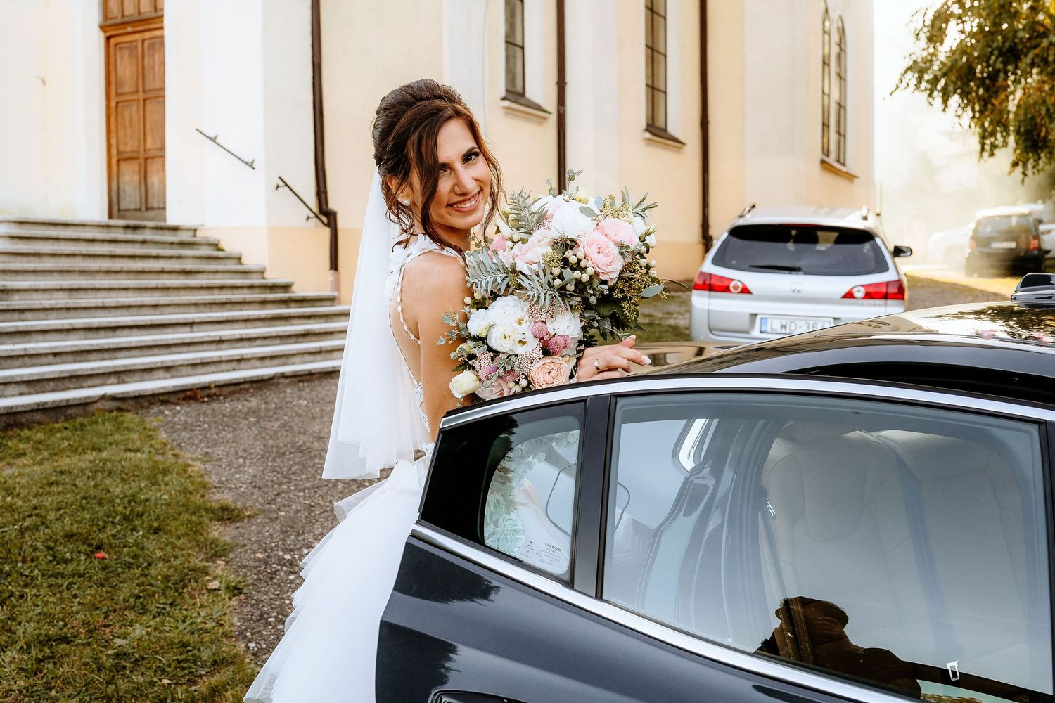 Bride with bouquet stands by car outside church stairs.