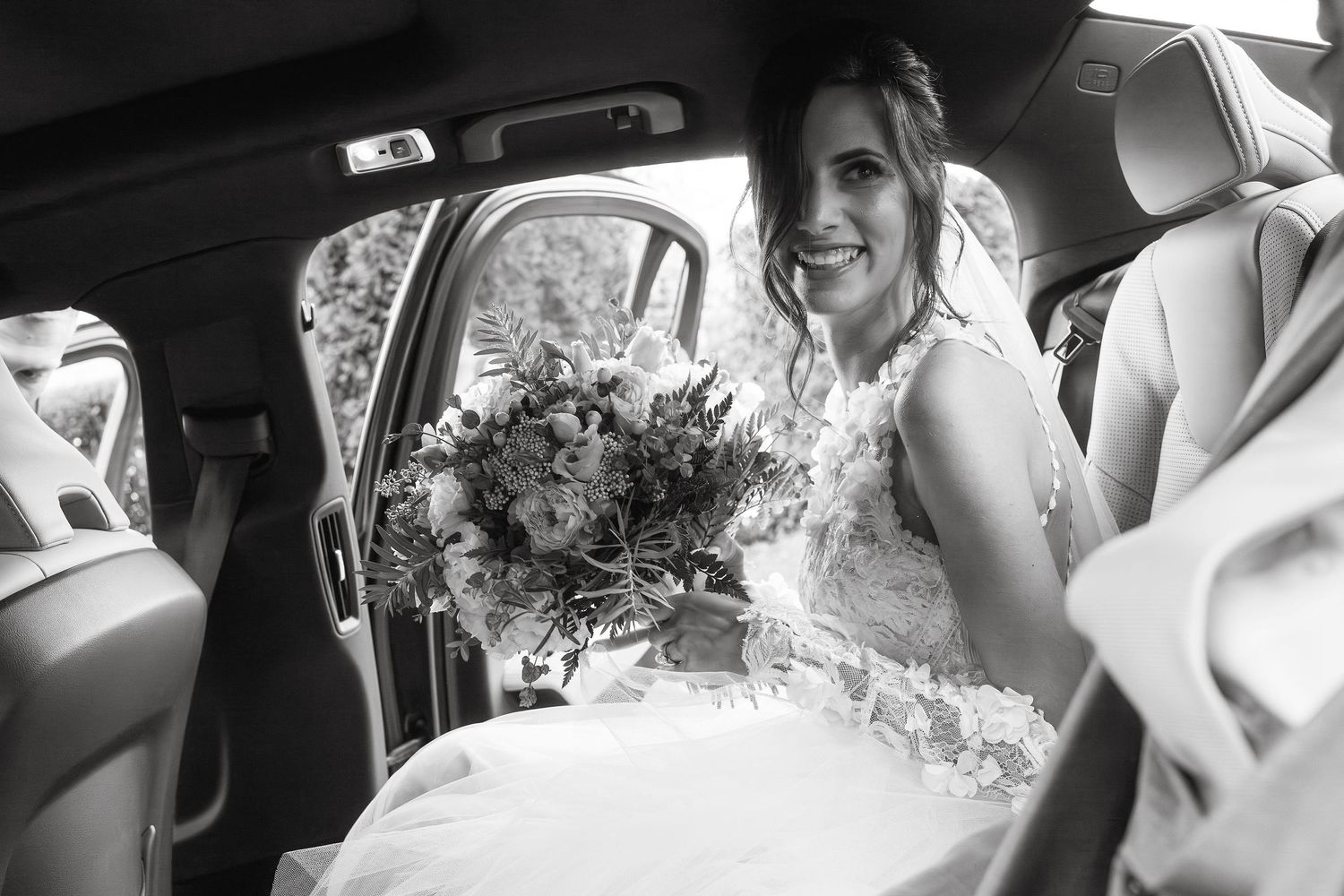 Black and white photo of bride sitting in car with wedding bouquet.