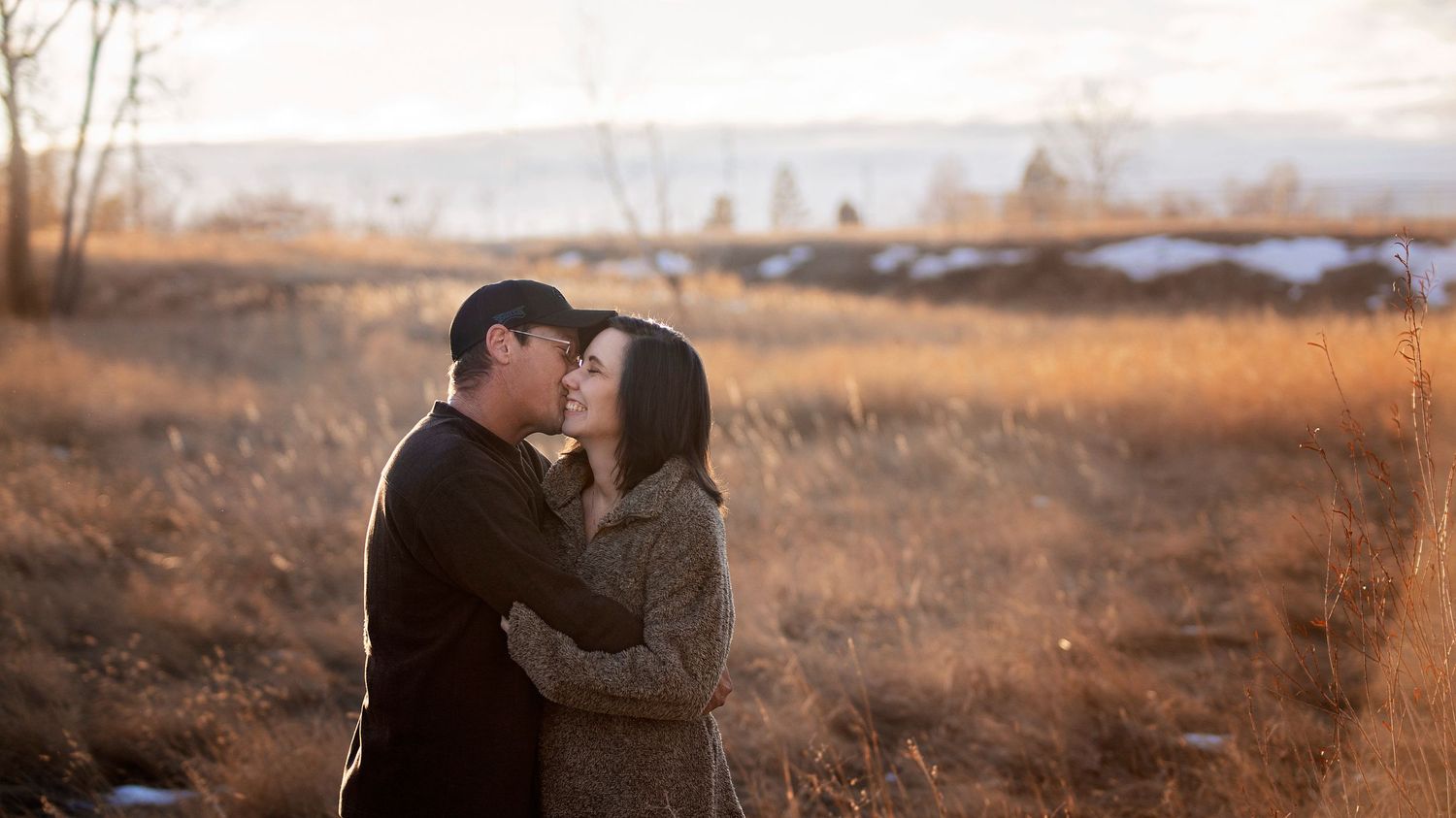 A couple shares an intimate moment together at sunset in a rustic field with mountains in the distance.