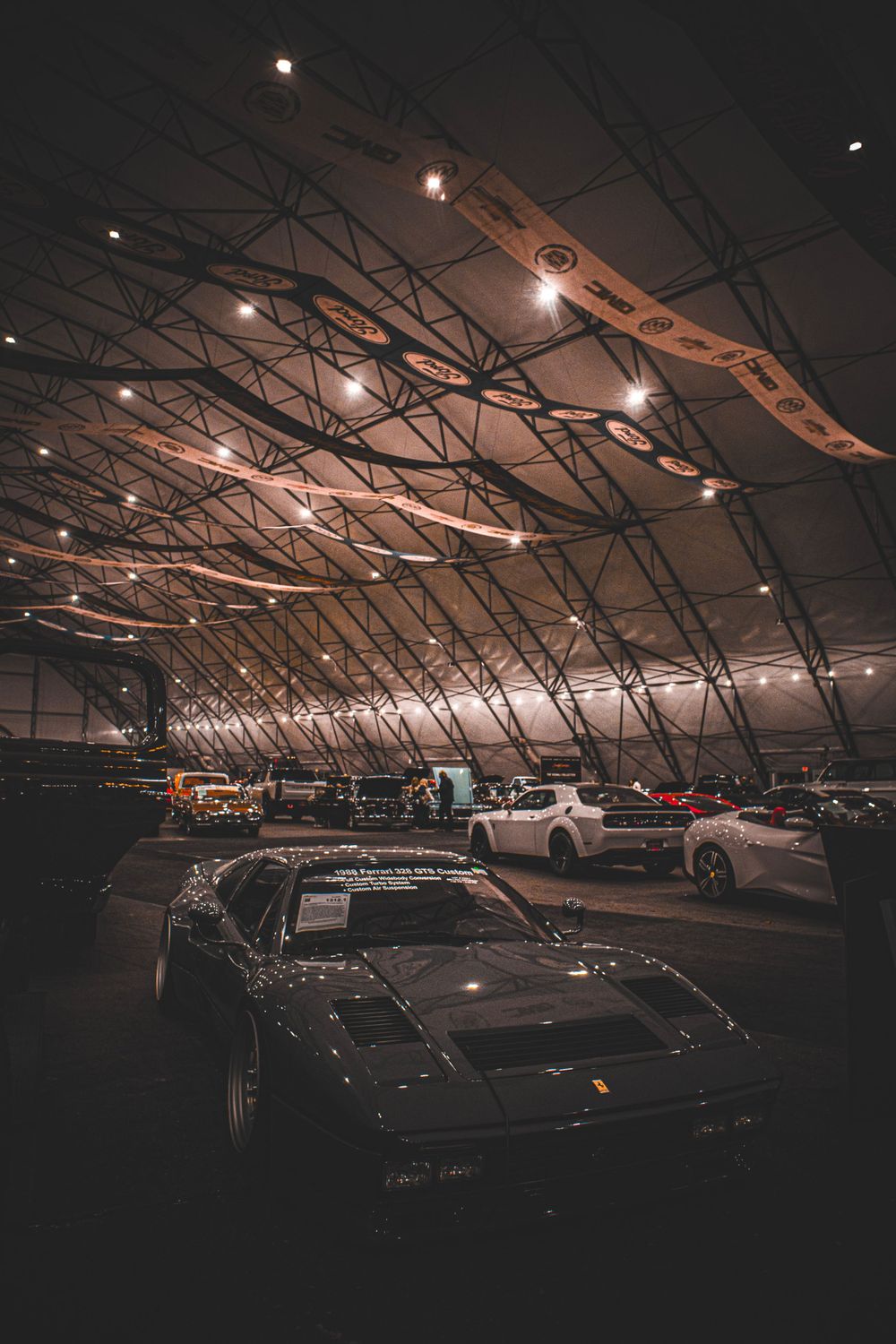 Car show venue with industrial ceiling and dramatic lighting showcasing multiple classic vehicles.