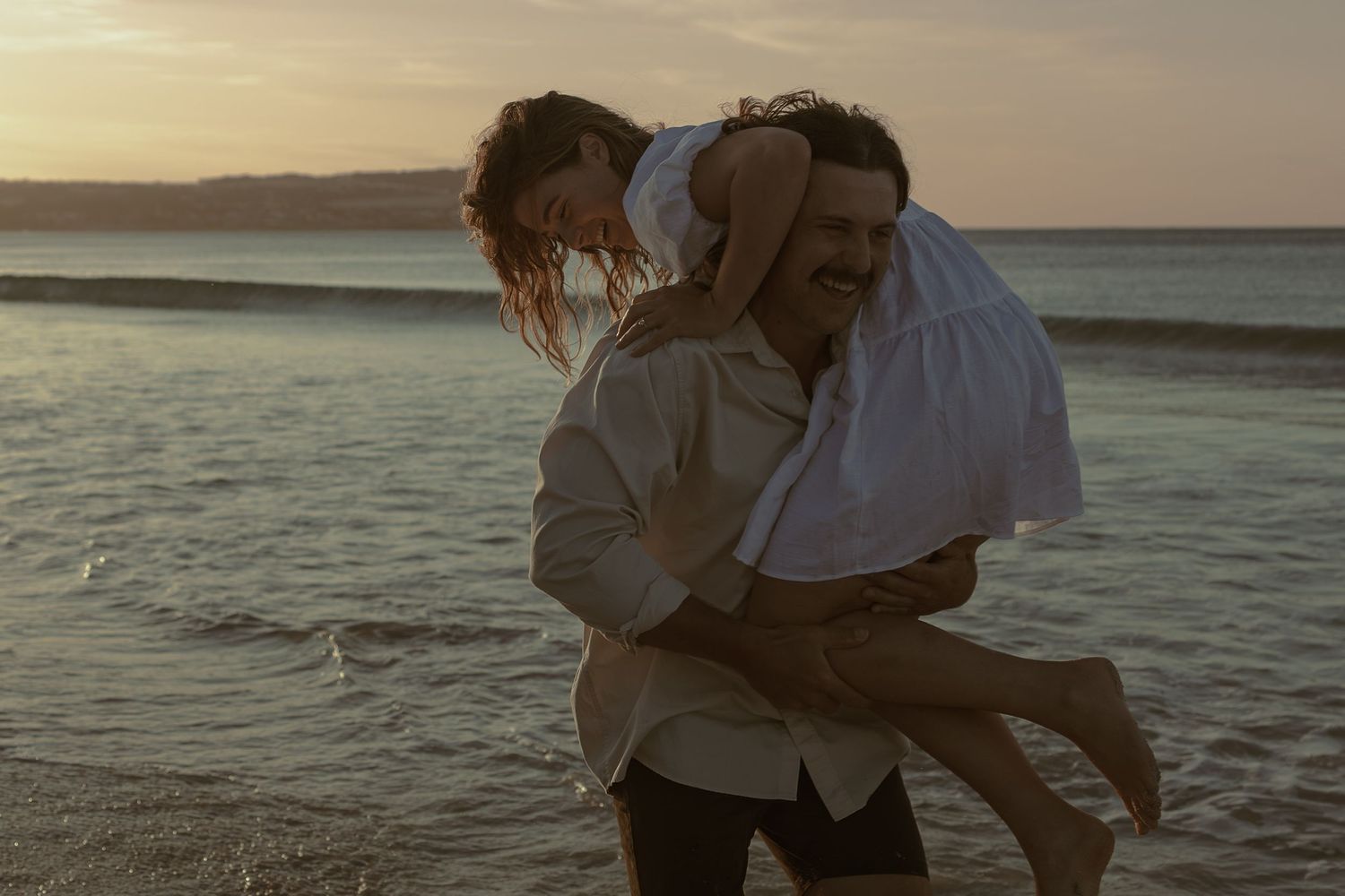 Romantic couple embracing on beach at sunset with golden light reflecting off ocean waves.