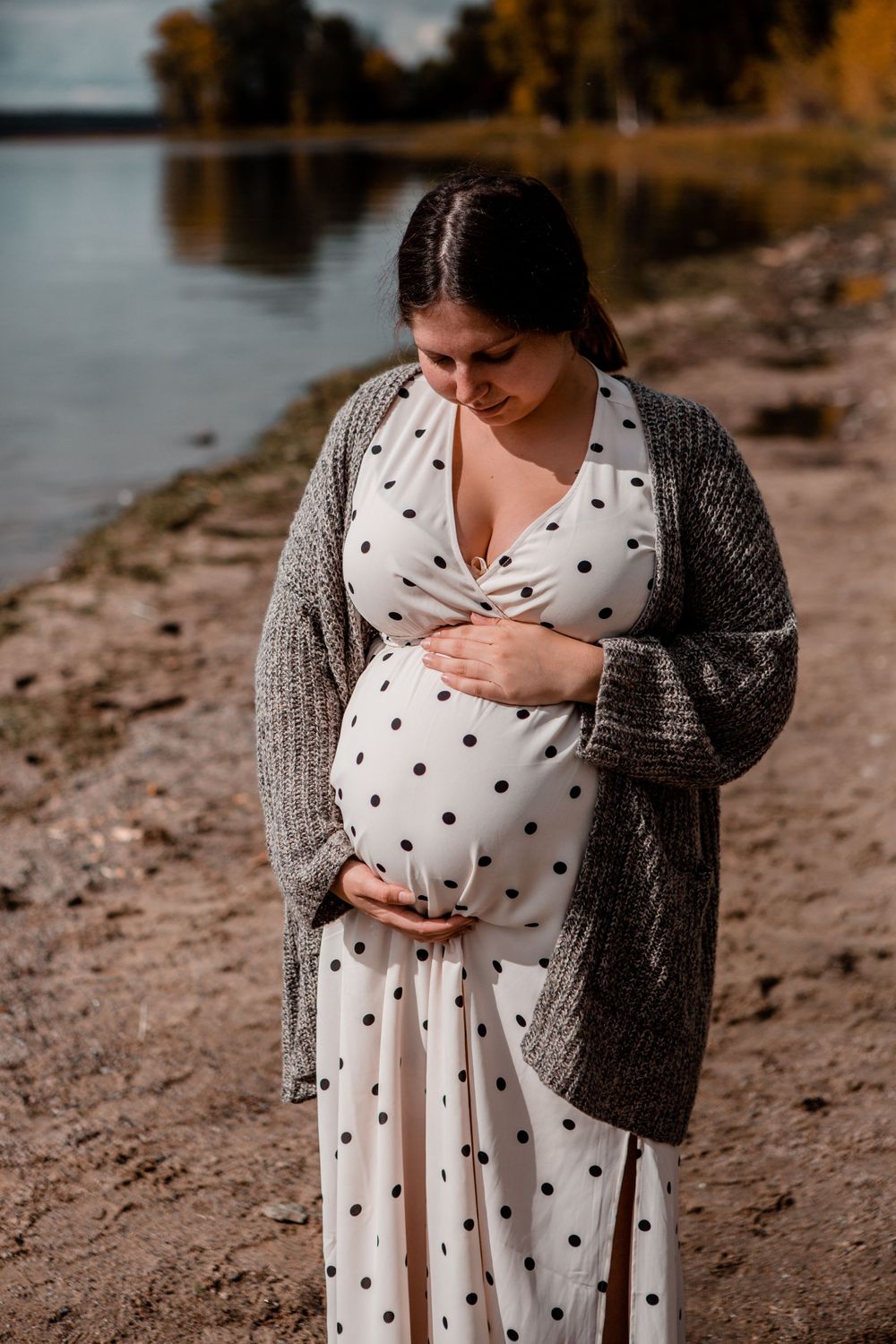 Expectant mother in polka dot dress and gray cardigan stands by lake during sunset.