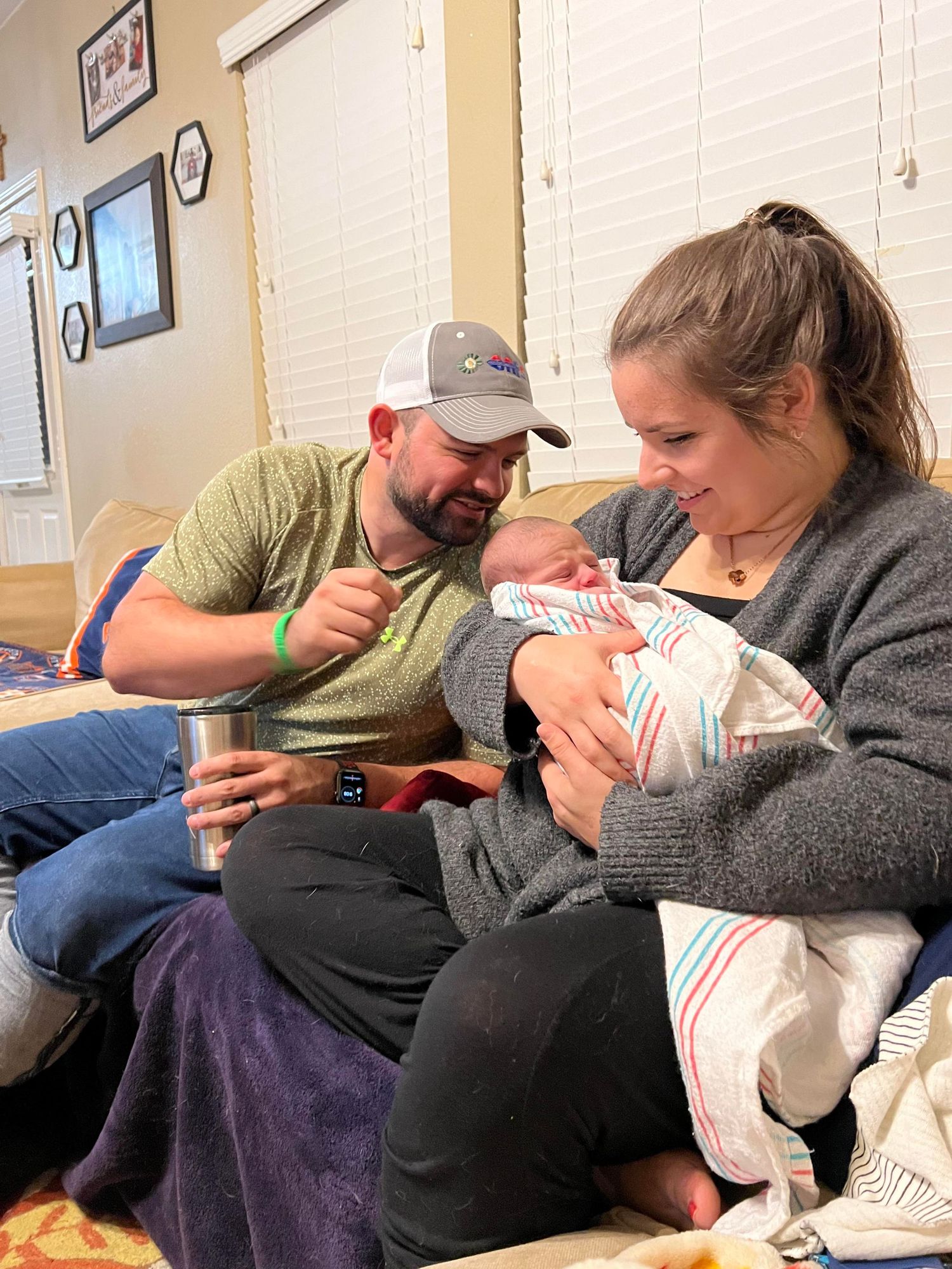 a man leans towards his wife who is wearing a grey shirt and holding a newborn baby wrapped in a striped blanket