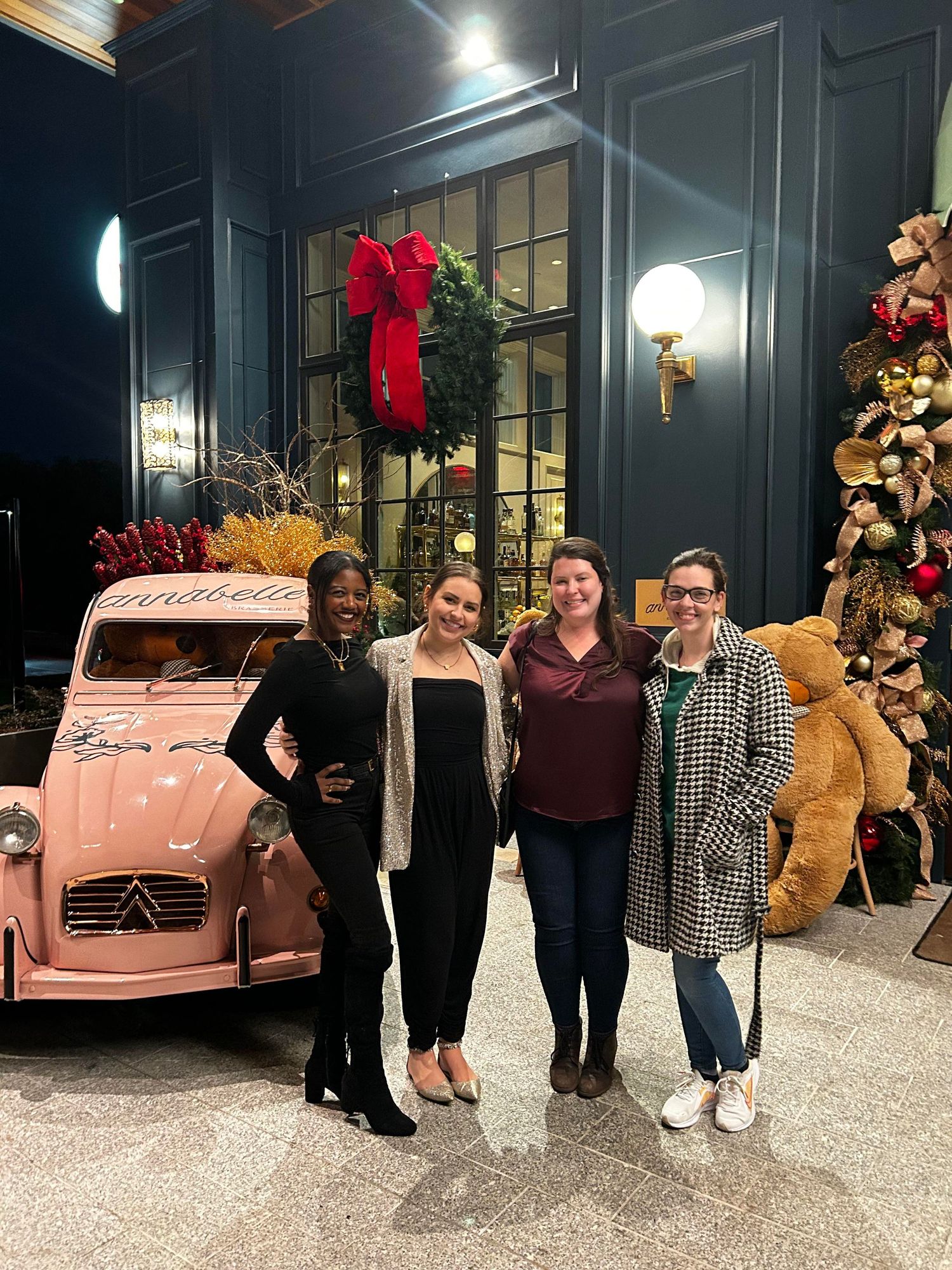 four women wearing winter wear stand in front of a pink classic car and multiple Christmas trees