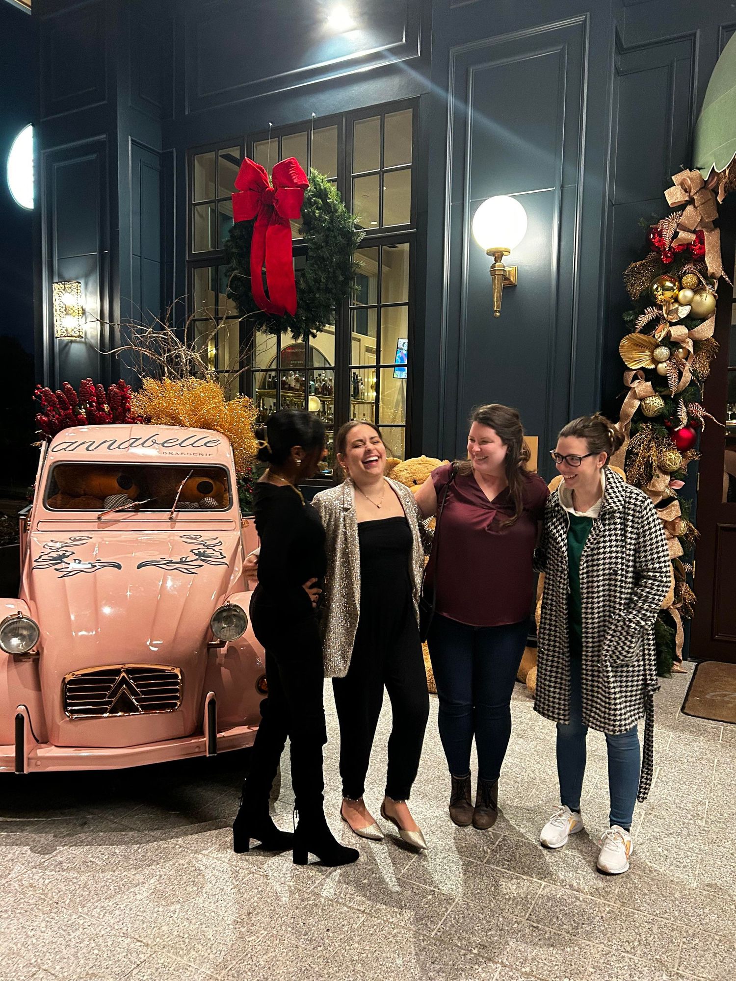 four women are posed in front of a pink classic car and multiple Christmas trees while laughing together